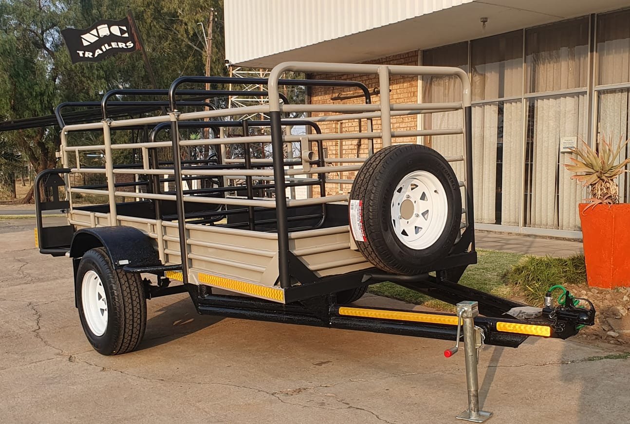 A flatbed utility trailer parked outside a building with large windows, with a spare tire mounted on the front corner and a flag advertising 'NIC Trailers' in the background.