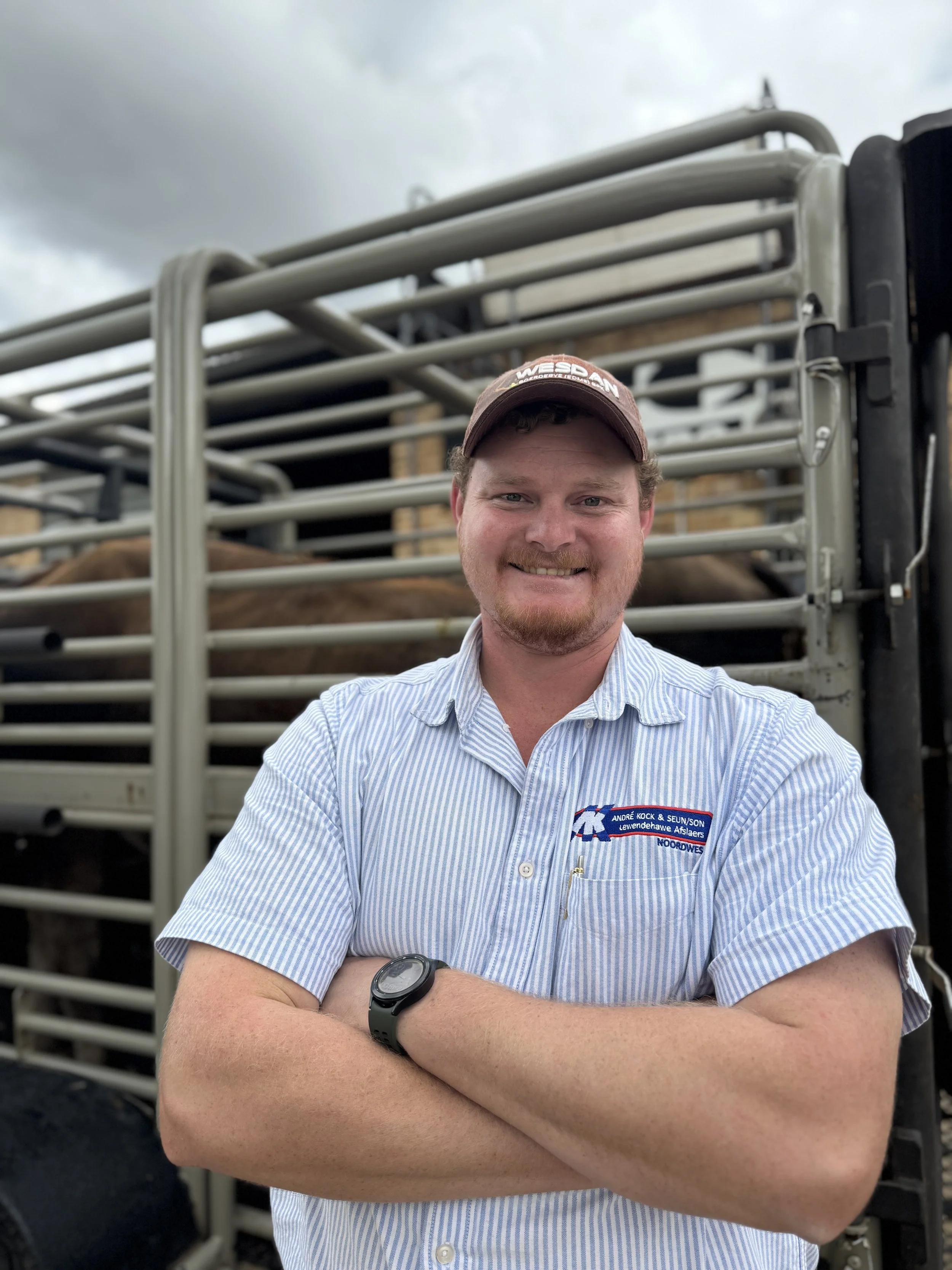 A man with a beard and short hair smiling with arms crossed, standing in front of a cattle truck, wearing a striped short-sleeve shirt with a company logo and a brown cap.