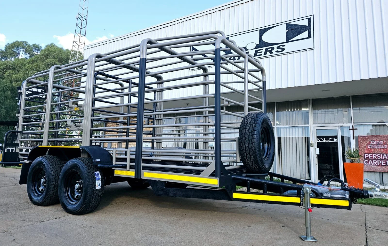 Metal livestock trailer with dual axles and spare tire parked outside a building with a sign that reads 'Neco.'