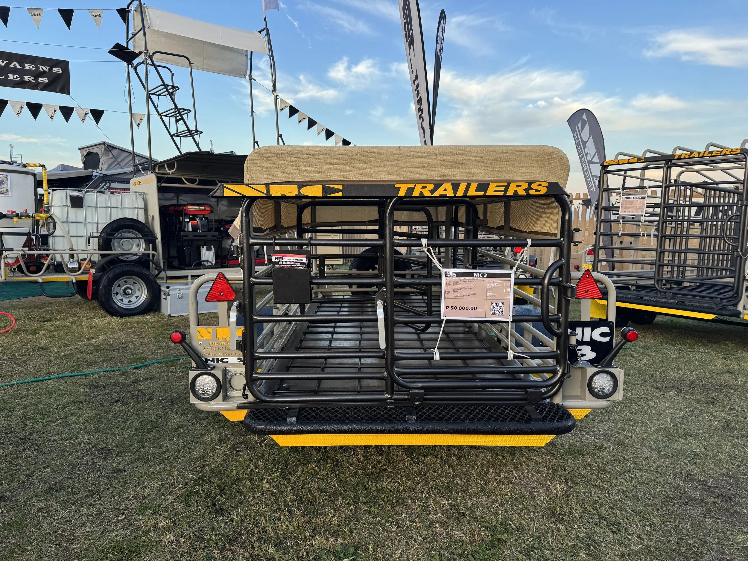 Display of a livestock trailer with a price tag at an outdoor equipment show. The trailer has a black metal frame and a canopy, with other trailers and equipment visible in the background under a blue sky.