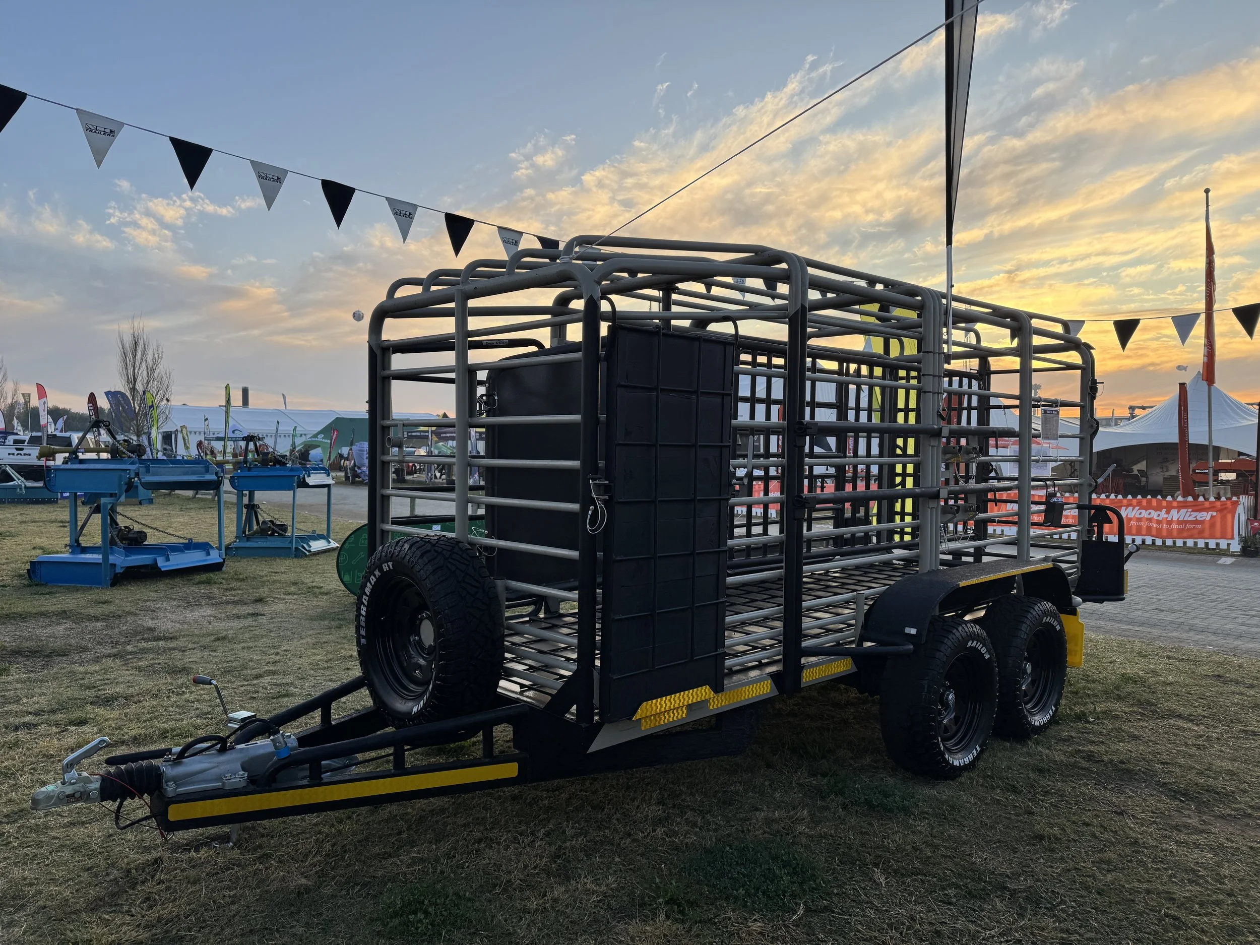Metal livestock trailer with dual axles and black tires parked on grass at outdoor event during sunset, with flags and tents in the background.