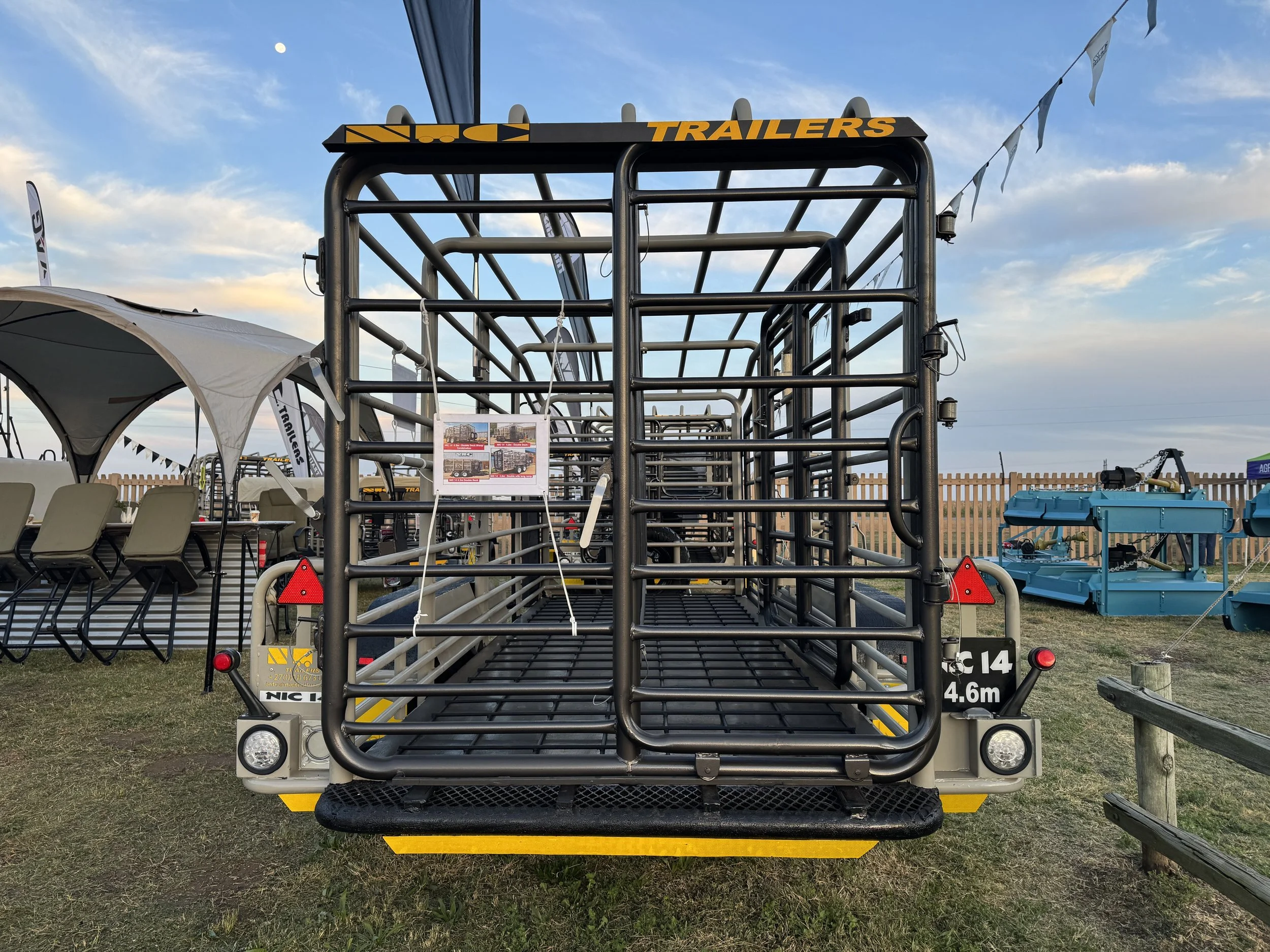 Black metal livestock trailer with yellow accents parked on grass with other blue trailers and outdoor tent in the background, under a partly cloudy sky.