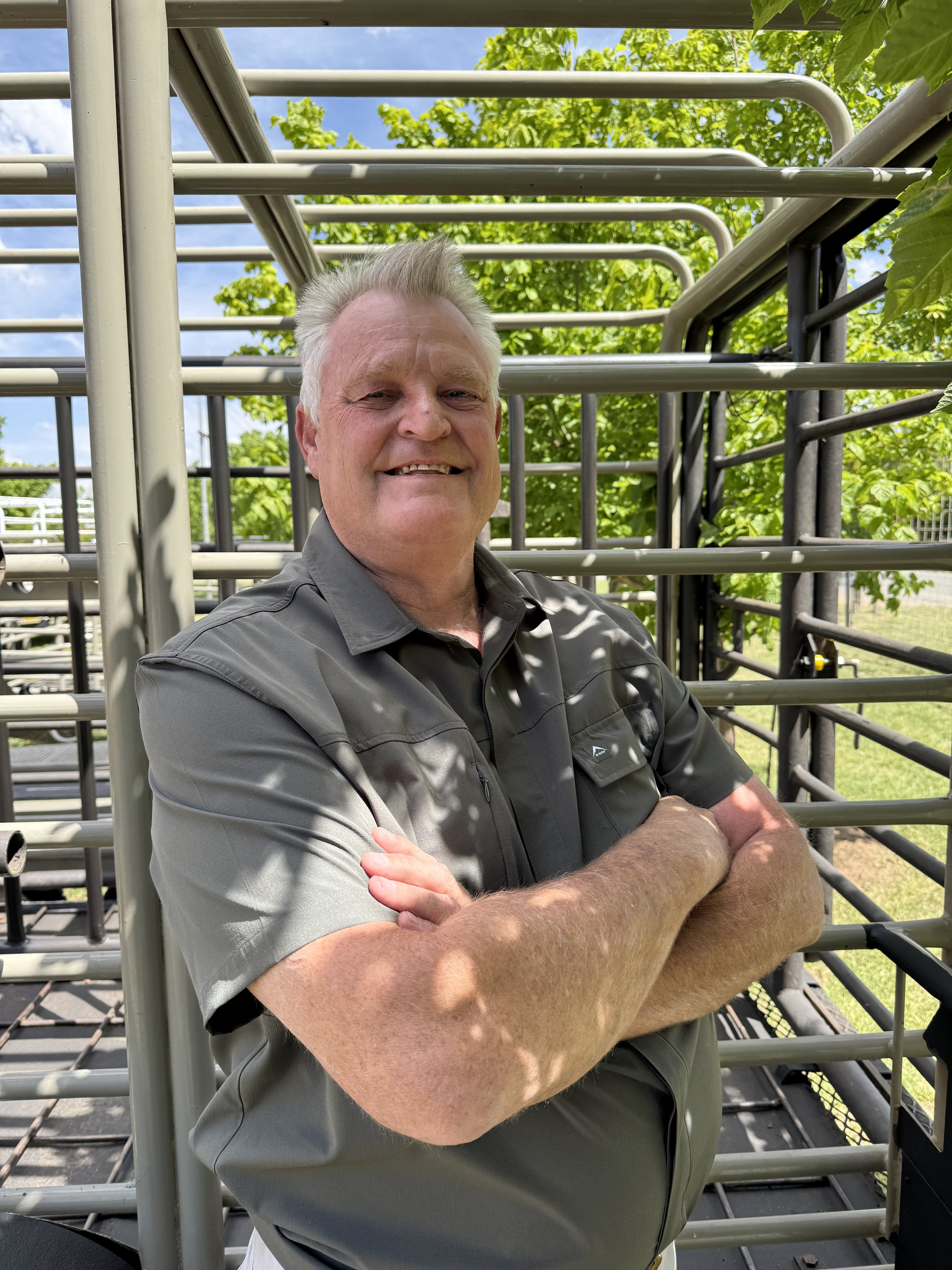 A smiling middle-aged man with gray hair standing outdoors inside a metal livestock pen, with green trees and a blue sky in the background.