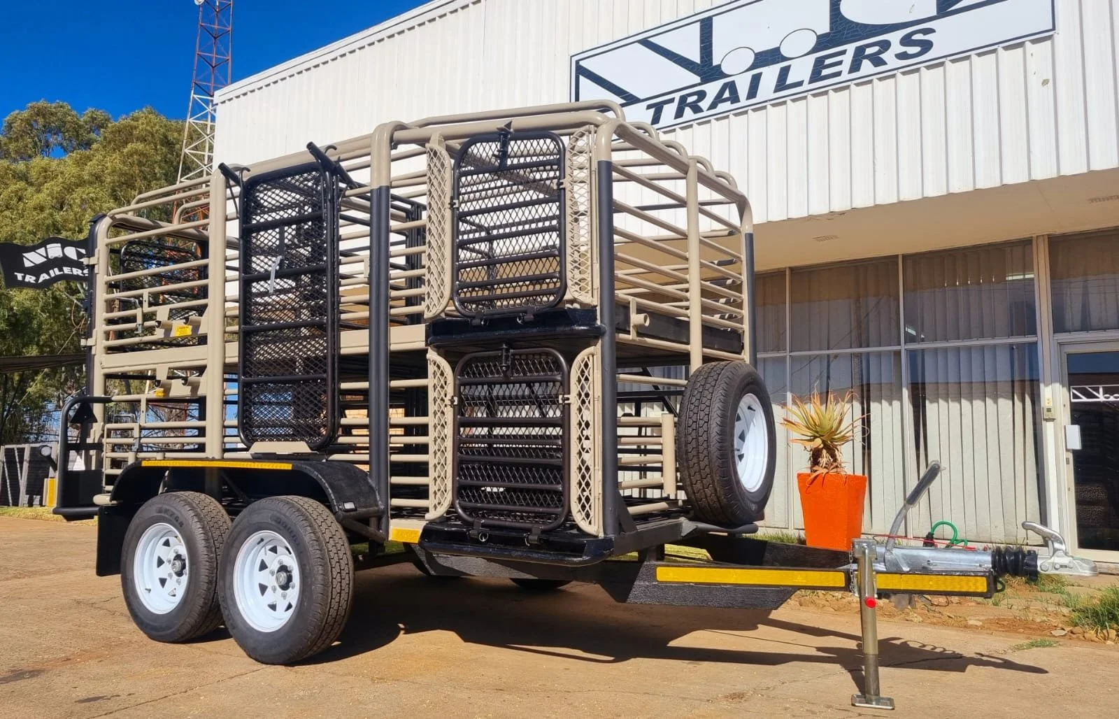 Livestock trailer with metal cage structure outside a building labeled "Trailers." Double axles and hitch visible, with a plant in an orange pot nearby.