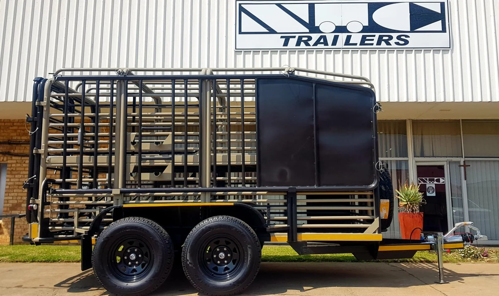 A livestock trailer parked outside a building with a sign that reads 'NCO Trailers', with a potted plant and some flowers nearby.