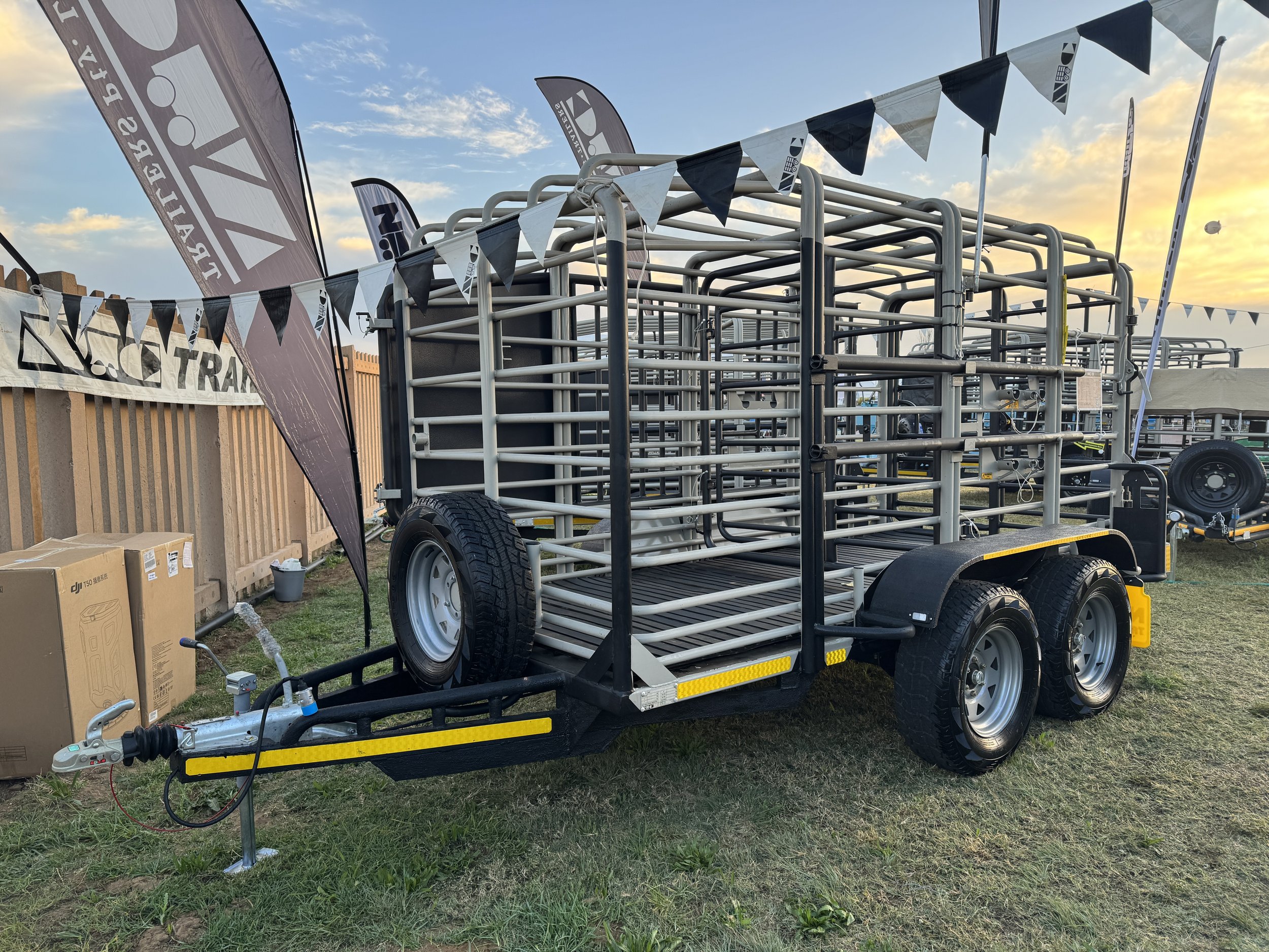 A metal livestock trailer with dual axles parked on grass at sunset, surrounded by flags and banners.