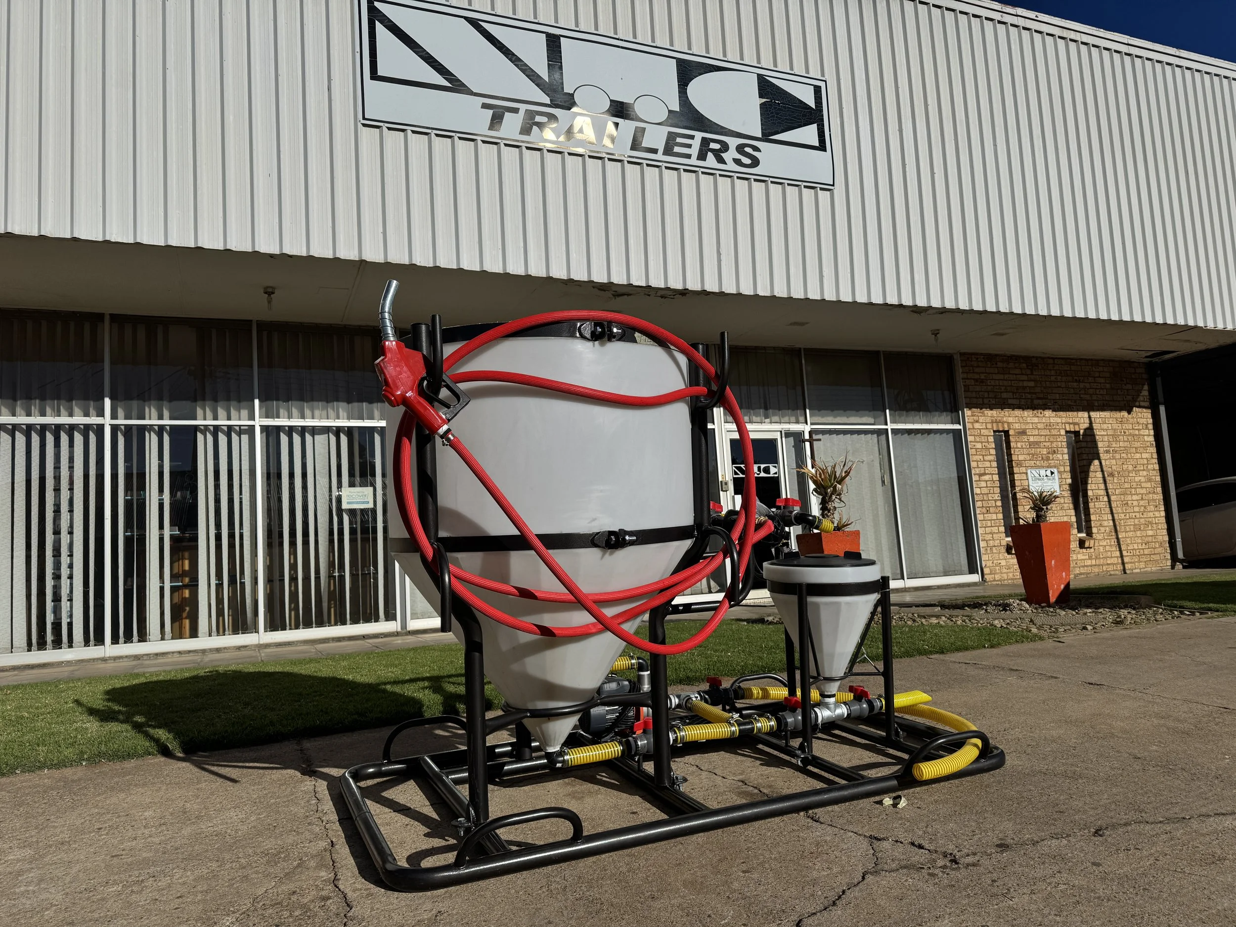 Large industrial sprayer with a white tank, red hoses, and yellow tubing, positioned outside a building with a sign that reads 'TRAILERS.'