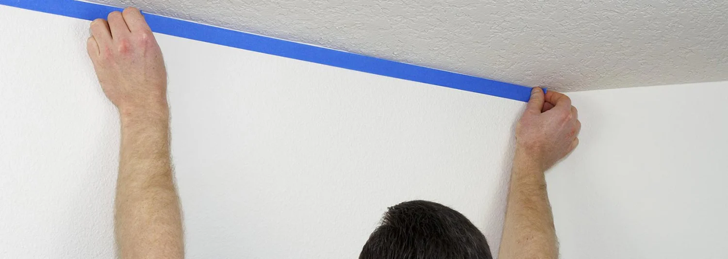 DIY painter in Silver Spring applying blue tape along a ceiling edge for crisp paint lines in a freshly prepped room.