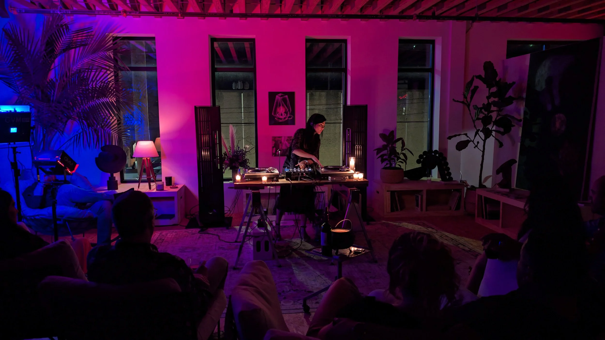 A woman DJing at an indoor event with purple and pink lighting, surrounded by plants and decor, with an audience watching.