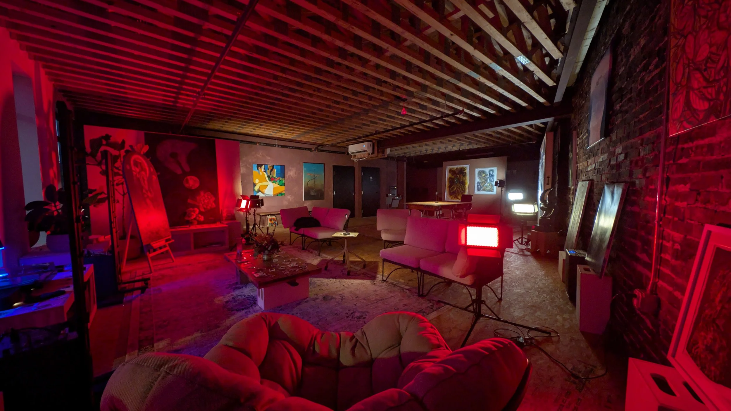 A dimly lit art gallery or studio space with various colorful paintings on the walls, red and blue lighting, and comfortable seating including sofa chairs and cushions, with exposed brick walls and wooden ceiling beams.