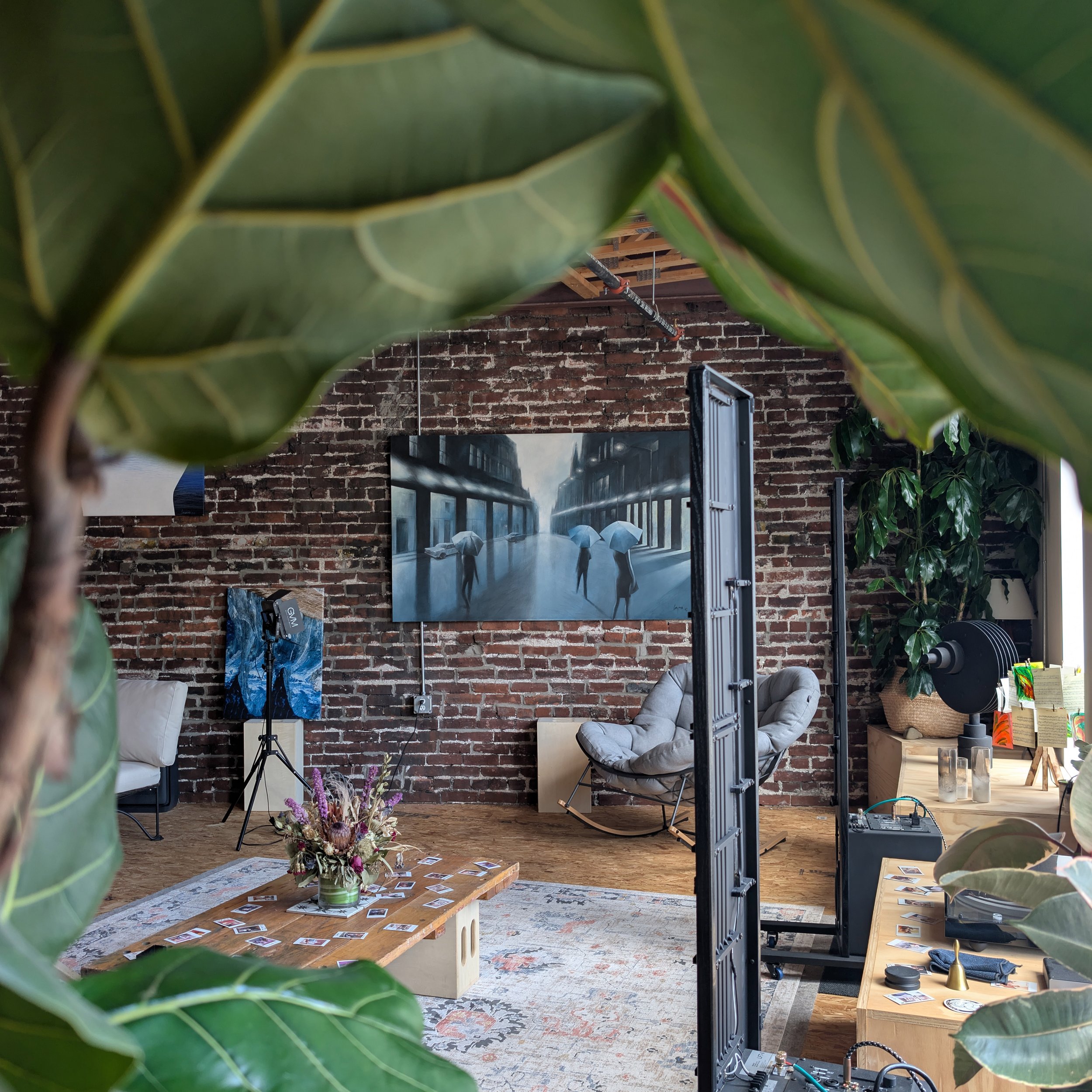 Interior of a modern living space with exposed brick wall, various paintings, a coffee table with photographs and flowers, a sofa, armchair, potted plants, and professional audio equipment, partially framed by large green leaves in the foreground.