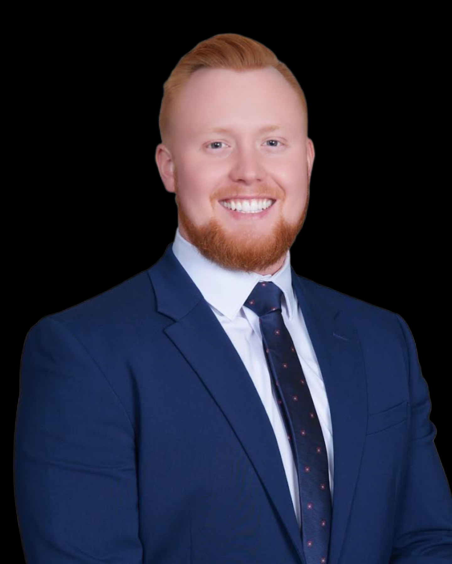 Headshot of a young man with red hair, beard, smiling, wearing a dark blue suit, white shirt, and dark tie, against a black background.