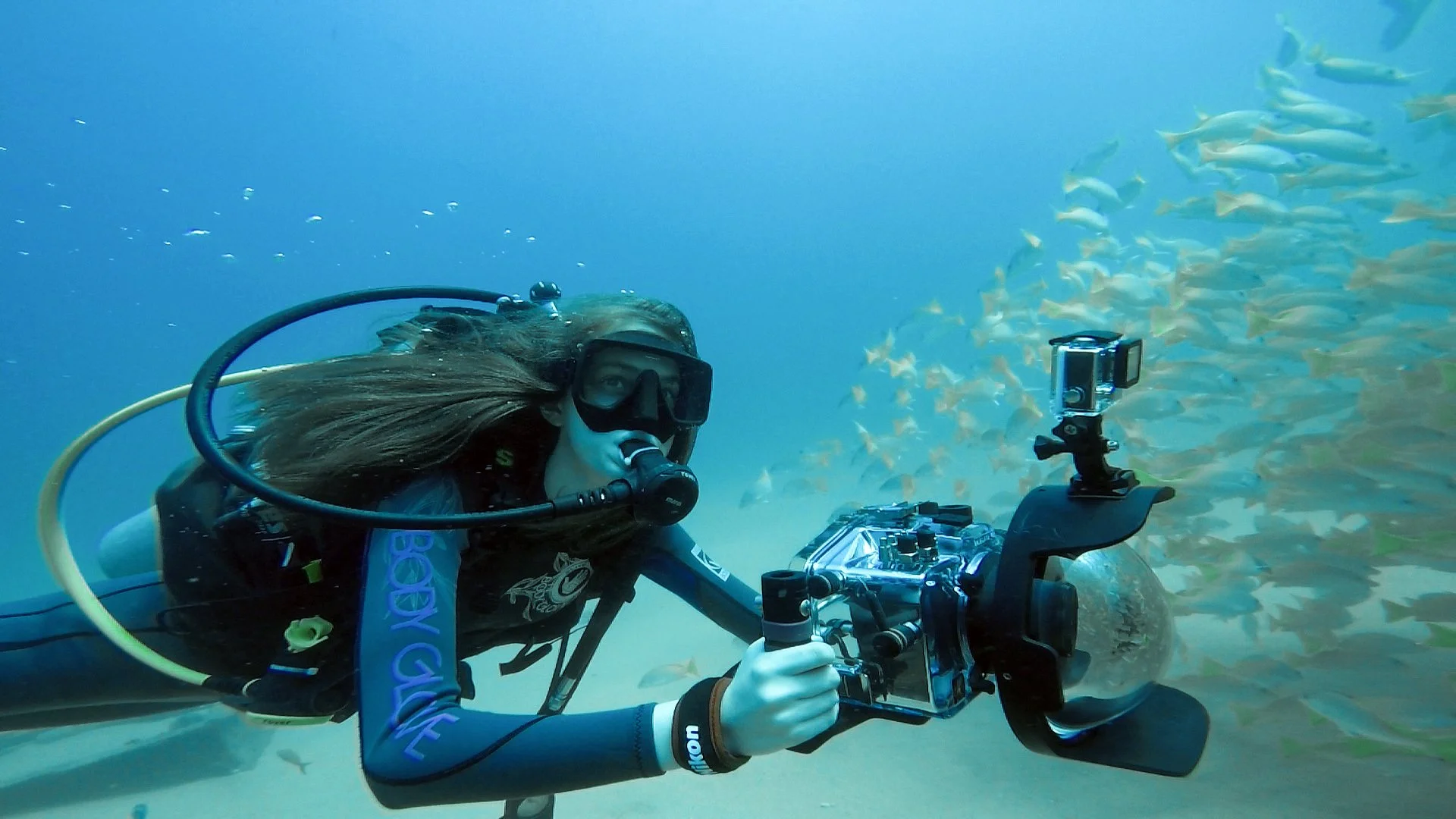 Person scuba diving underwater surrounded by a school of fish, holding a camera.