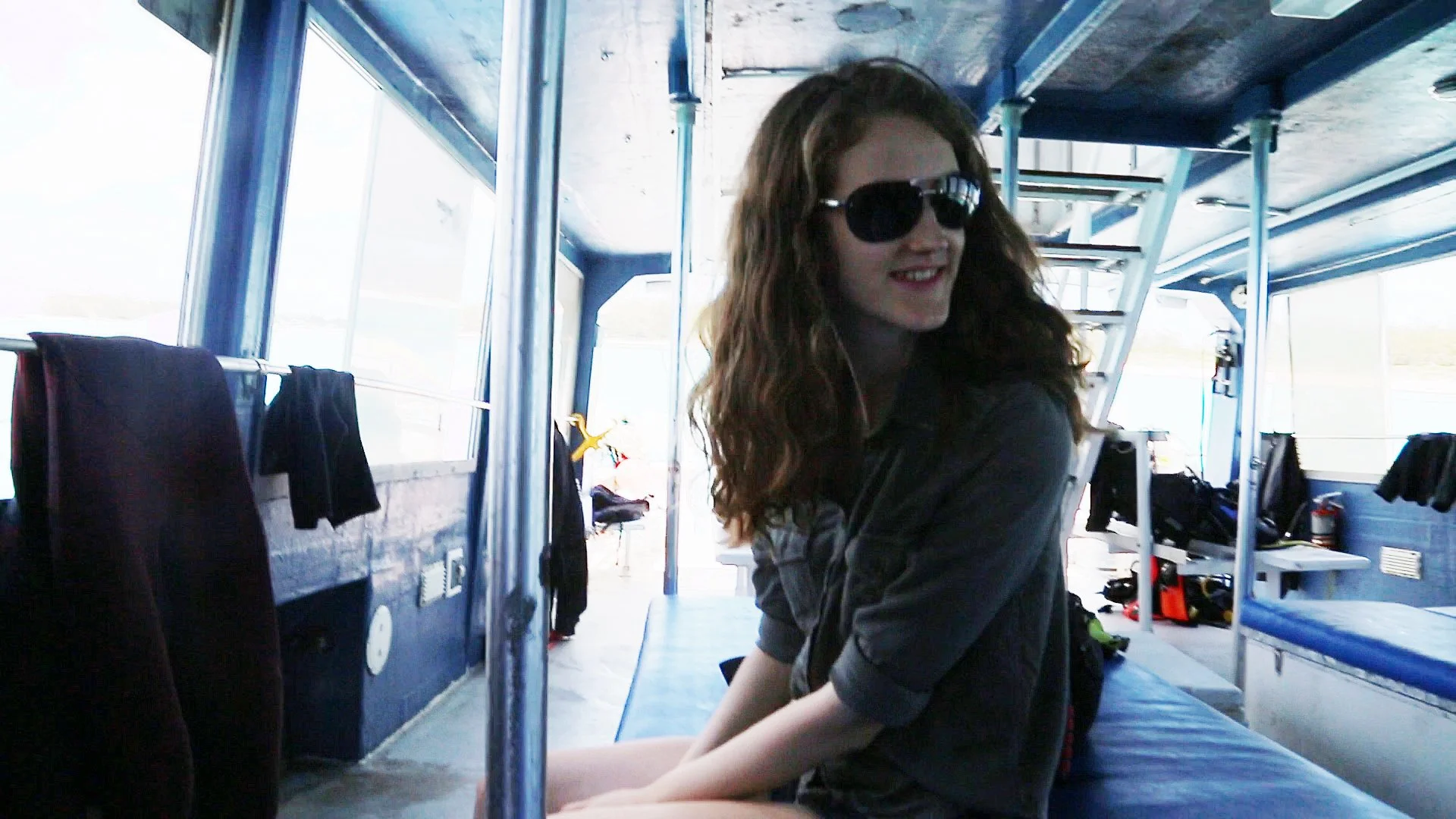 Young woman with curly hair wearing sunglasses and a green shirt, sitting on a bench inside a dive boat, smiling and looking back.