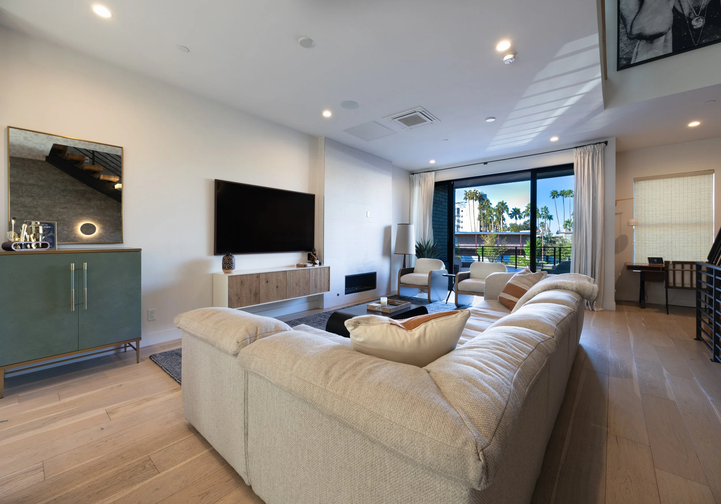 Modern living room with a large beige sofa, flat-screen TV on the wall, light wood flooring, two armchairs, and sliding glass doors leading to a balcony with palm trees visible outside.