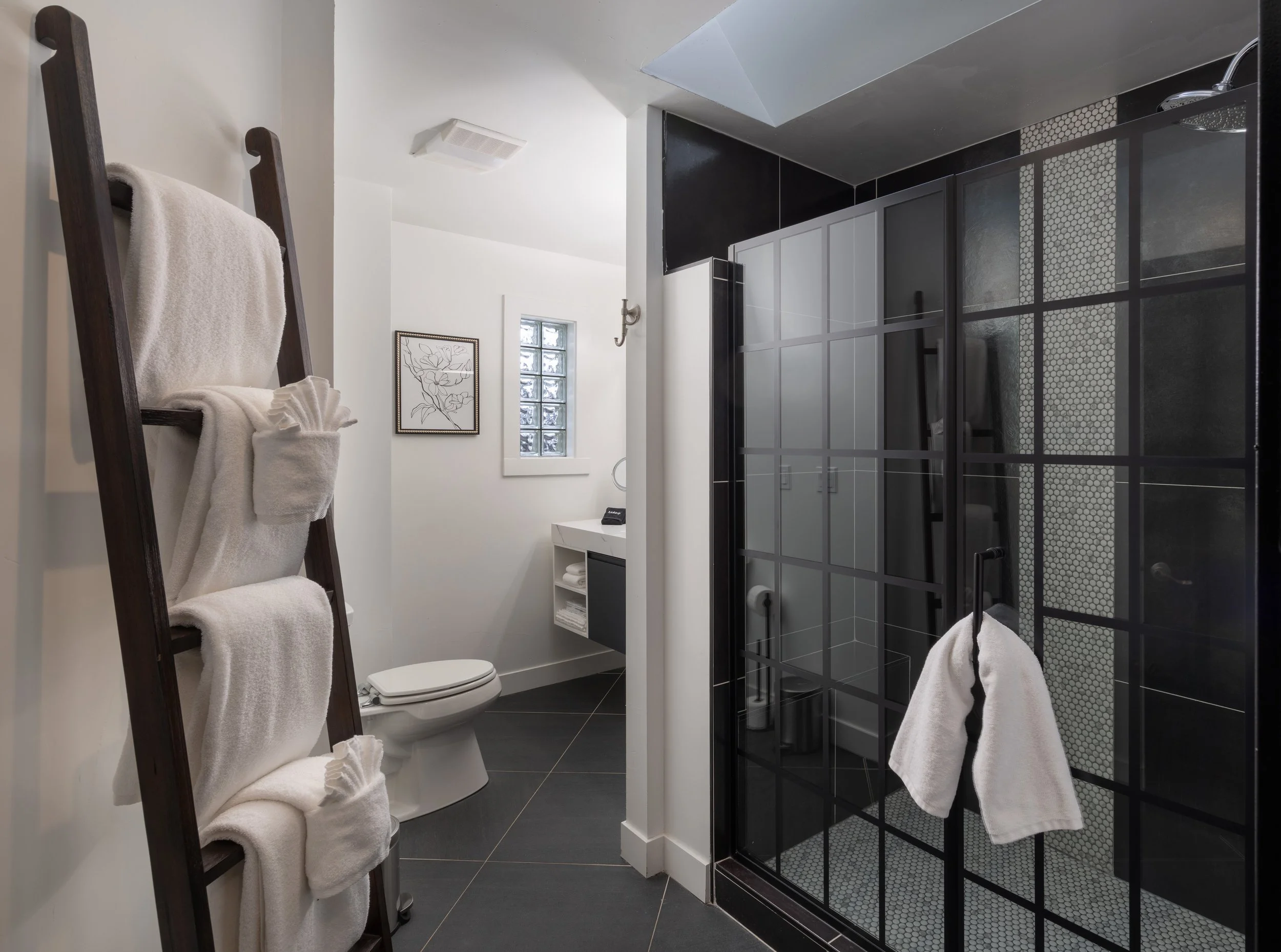 Modern bathroom with a black-framed glass shower, white towels on a ladder rack, and a white sink. Black floor tiles and a small window add contrast.