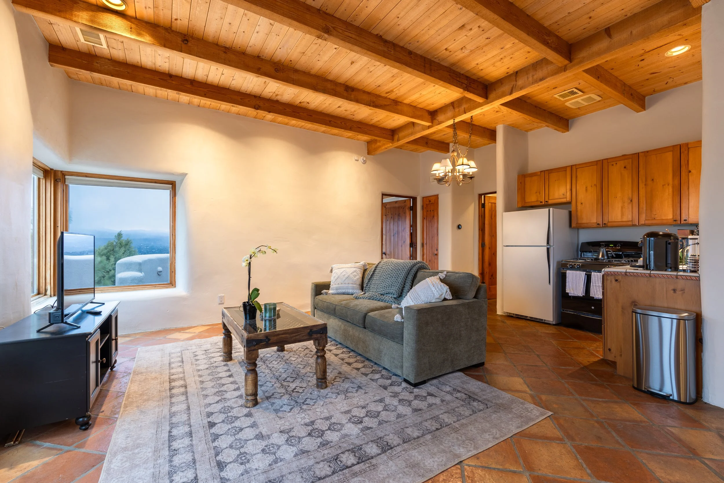 Cozy living room with wooden beams, sofa, TV, coffee table, kitchen area, and large window.