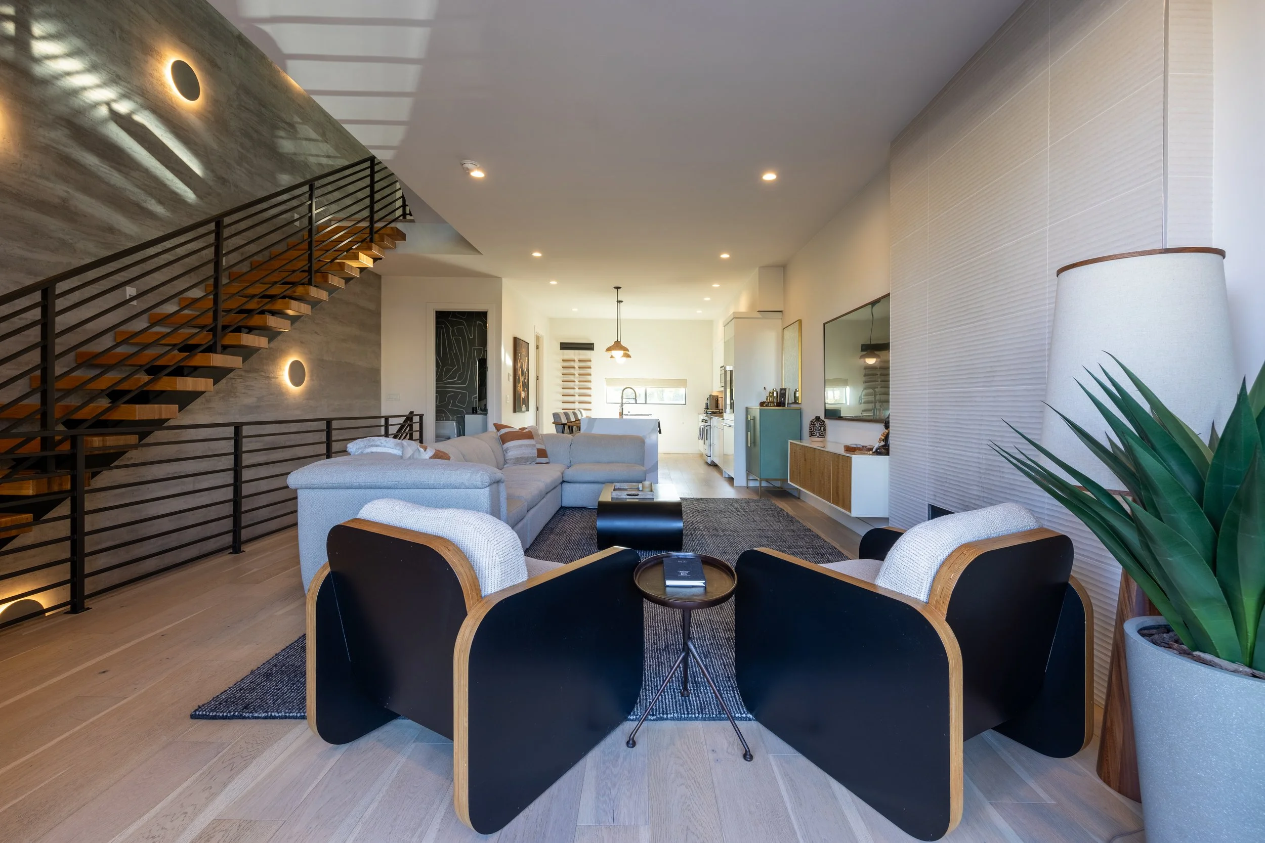 Modern living room with a staircase, gray sectional sofa, armchairs, and a large flat-screen TV. The space has wooden flooring, a floor lamp, and a potted plant.