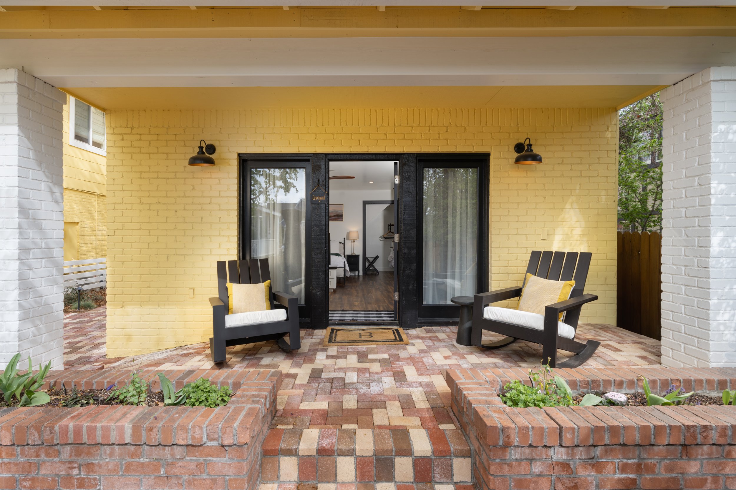 Front porch with two black Adirondack chairs, yellow pillows, brick flooring, and yellow painted brick wall with black framed glass door leading into a cozy interior.