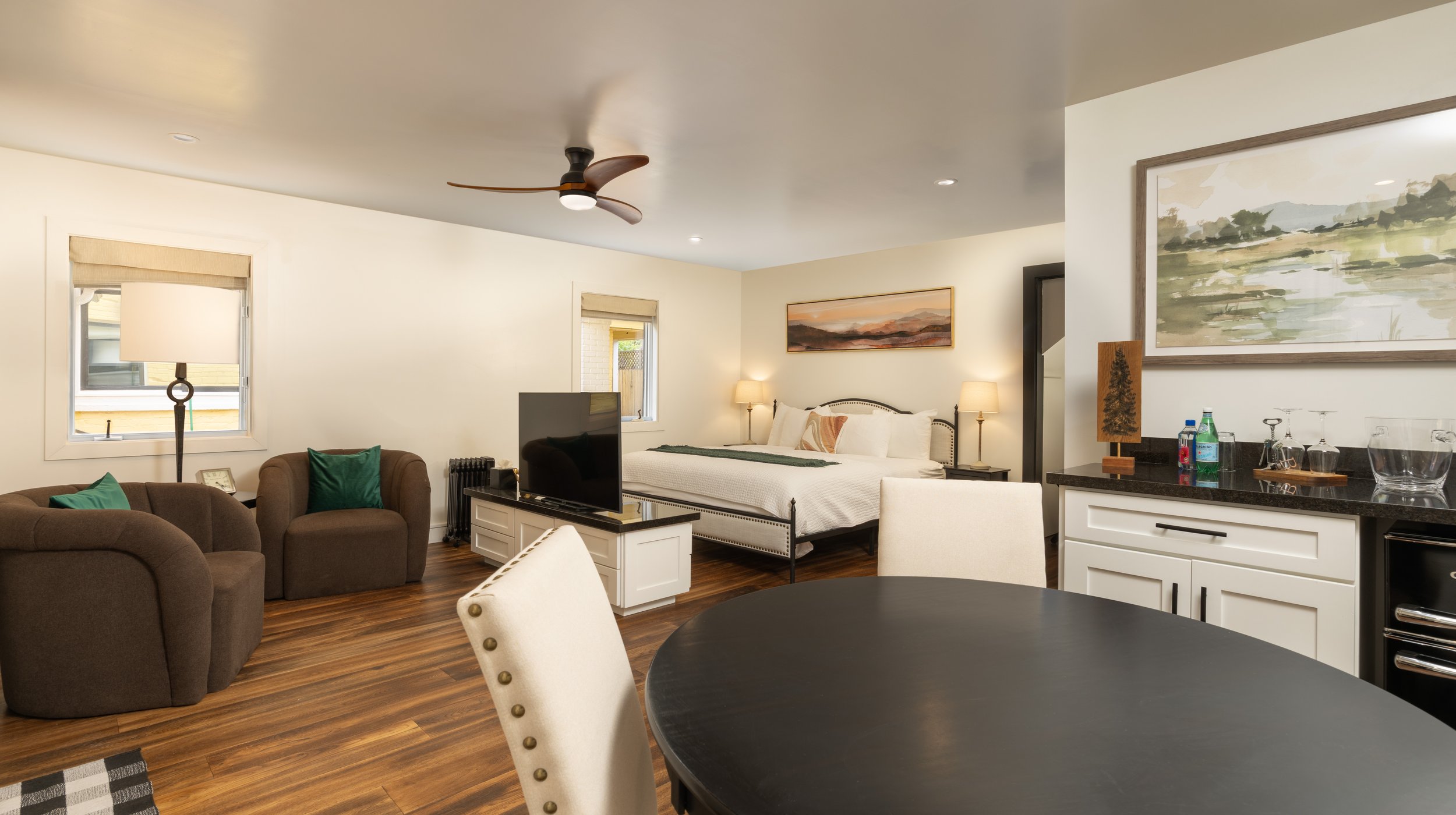 Modern studio apartment interior with a bed, armchairs, TV, and kitchen area. The room features wooden flooring, a ceiling fan, and minimalist decor.