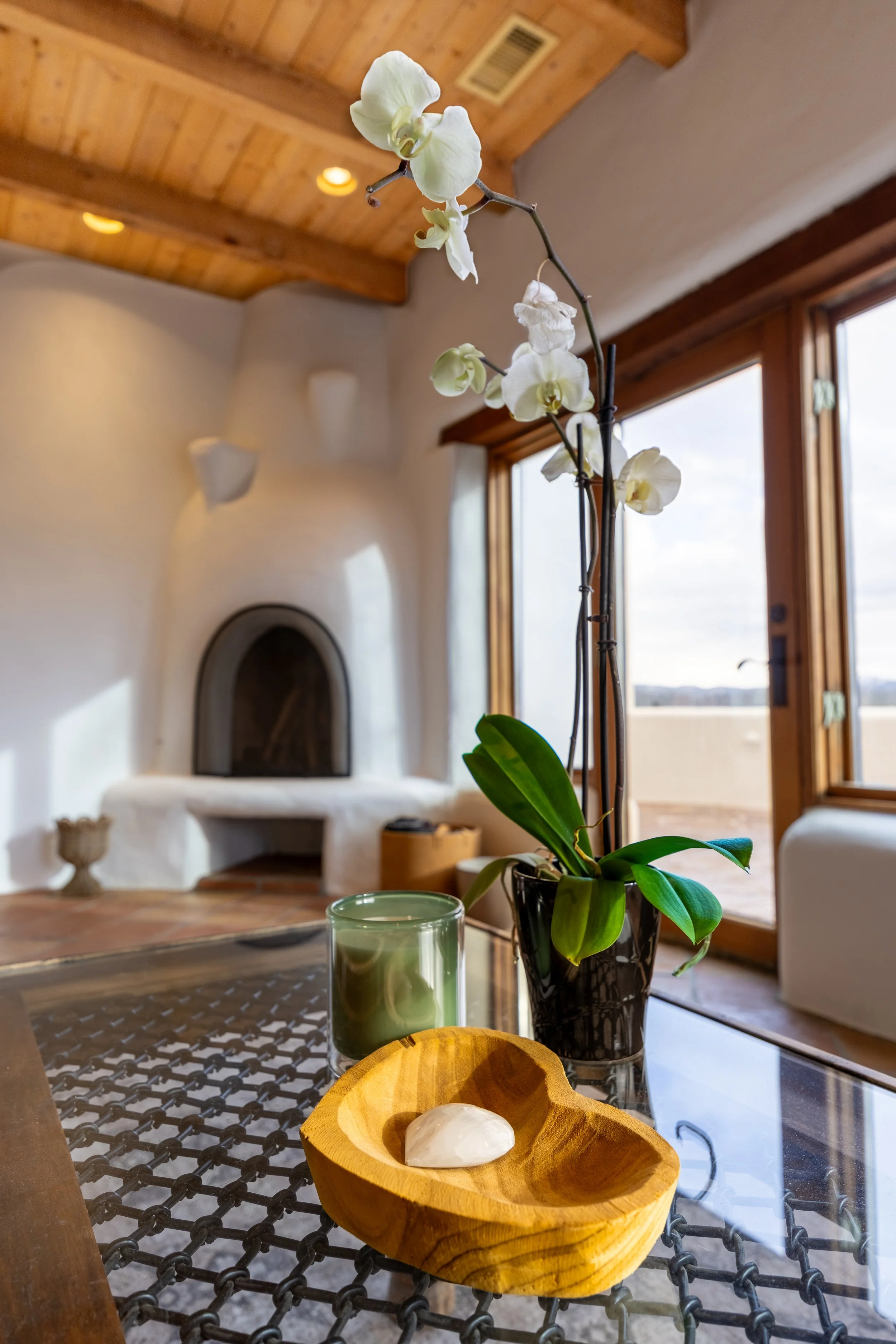 Interior of a modern room featuring a white orchid in a black pot on a glass table, a green candle, a wooden bowl, a white fireplace with a rounded design, and large windows with wooden frames.