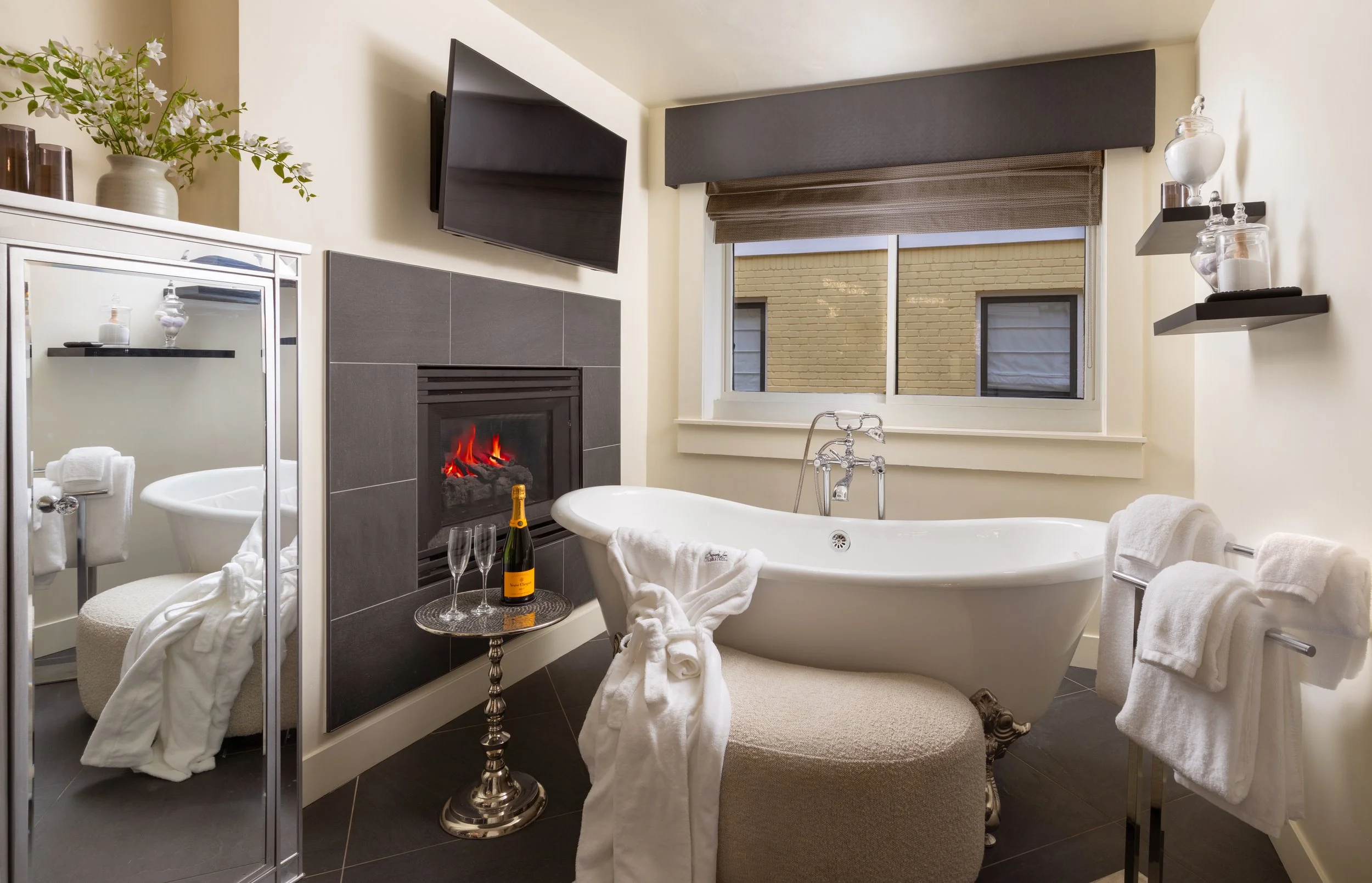Luxurious bathroom with a clawfoot bathtub, champagne, fireplace, flat-screen TV, small mirror, and decorative items.