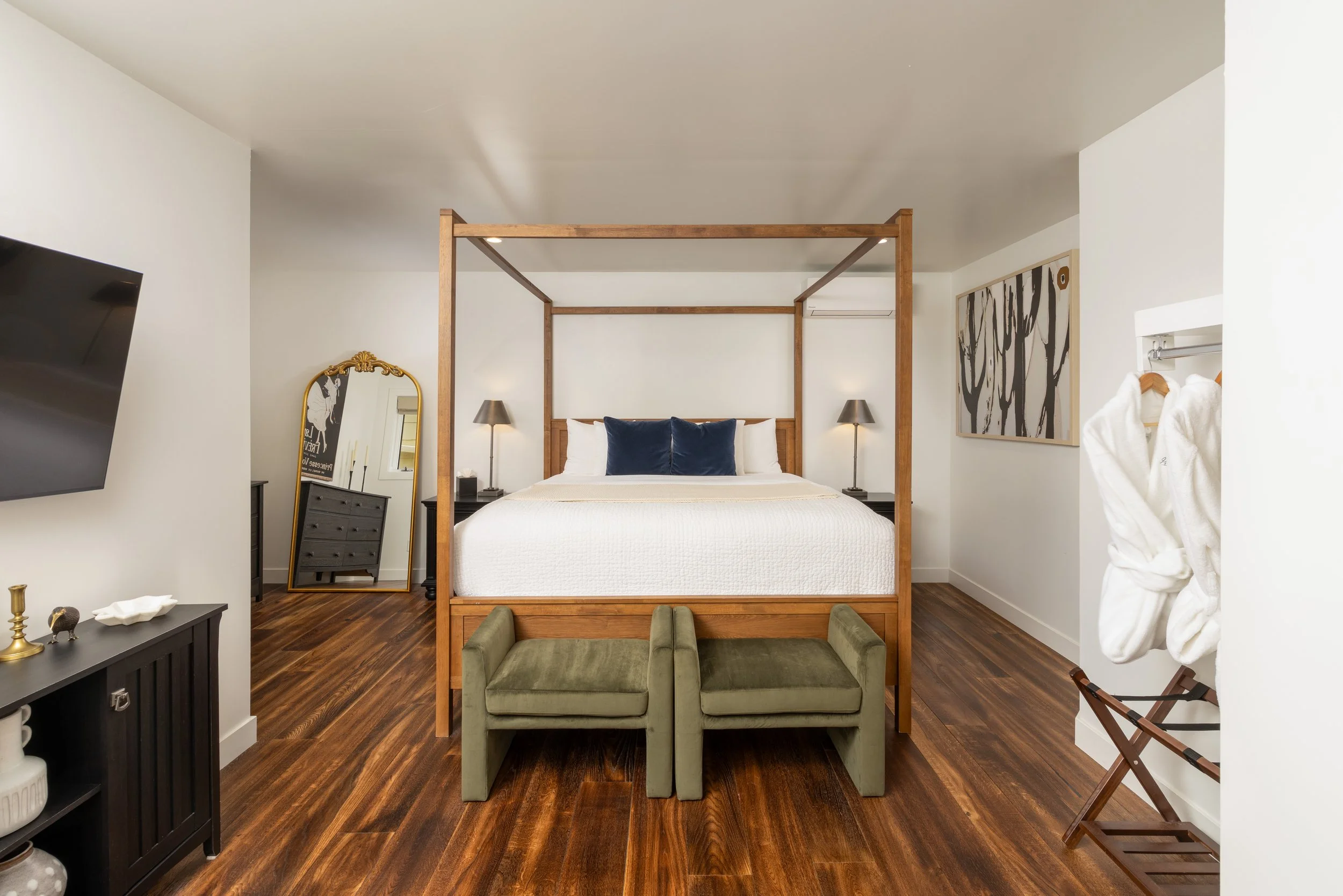 Contemporary bedroom with a four-poster bed, white bedding, two green benches, large mirror, TV, wooden floors, wall art, and a pair of white bathrobes on a hanger.