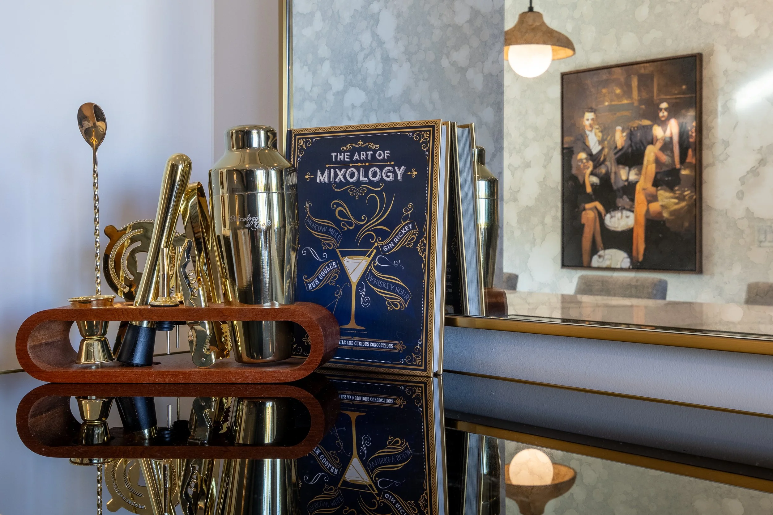 Mixology book and cocktail tools on mirrored surface, with a reflection of a painting in the background.