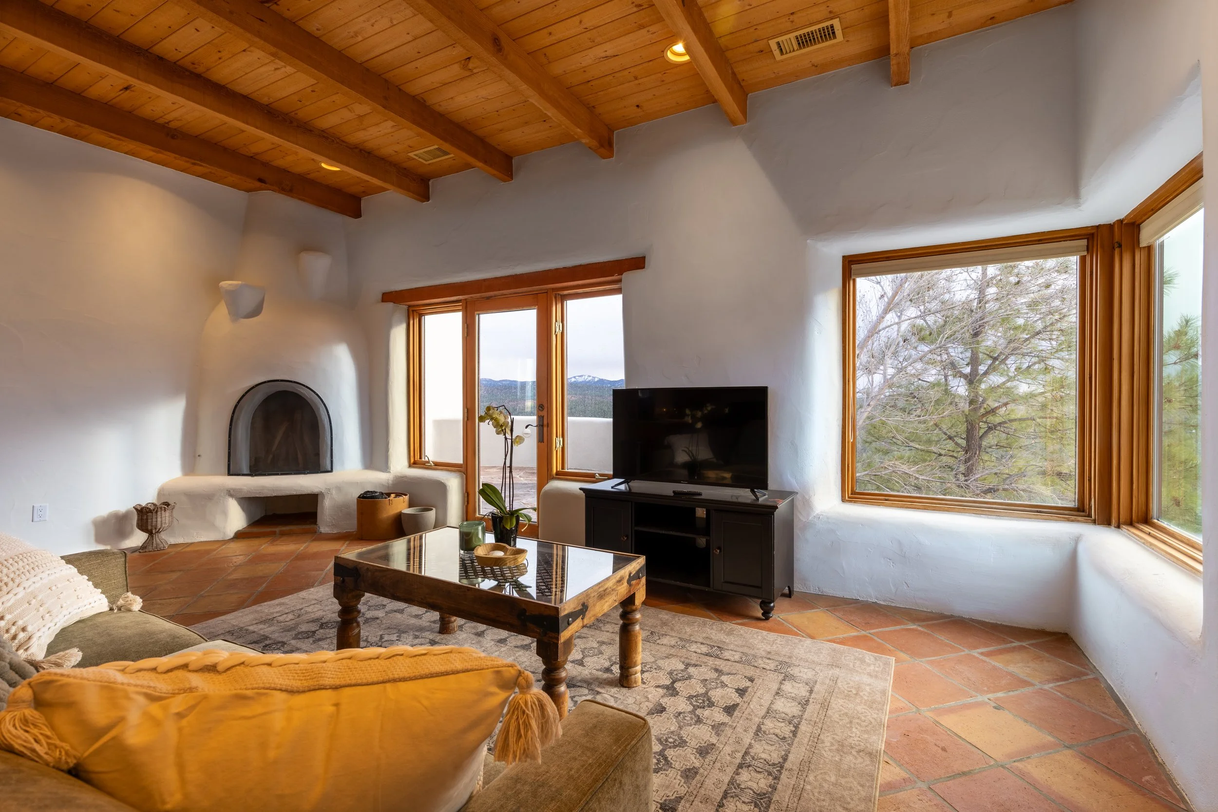 Southwestern style living room with terracotta tile flooring, a kiva fireplace, large windows overlooking a scenic view, wooden ceiling beams, and modern furniture.