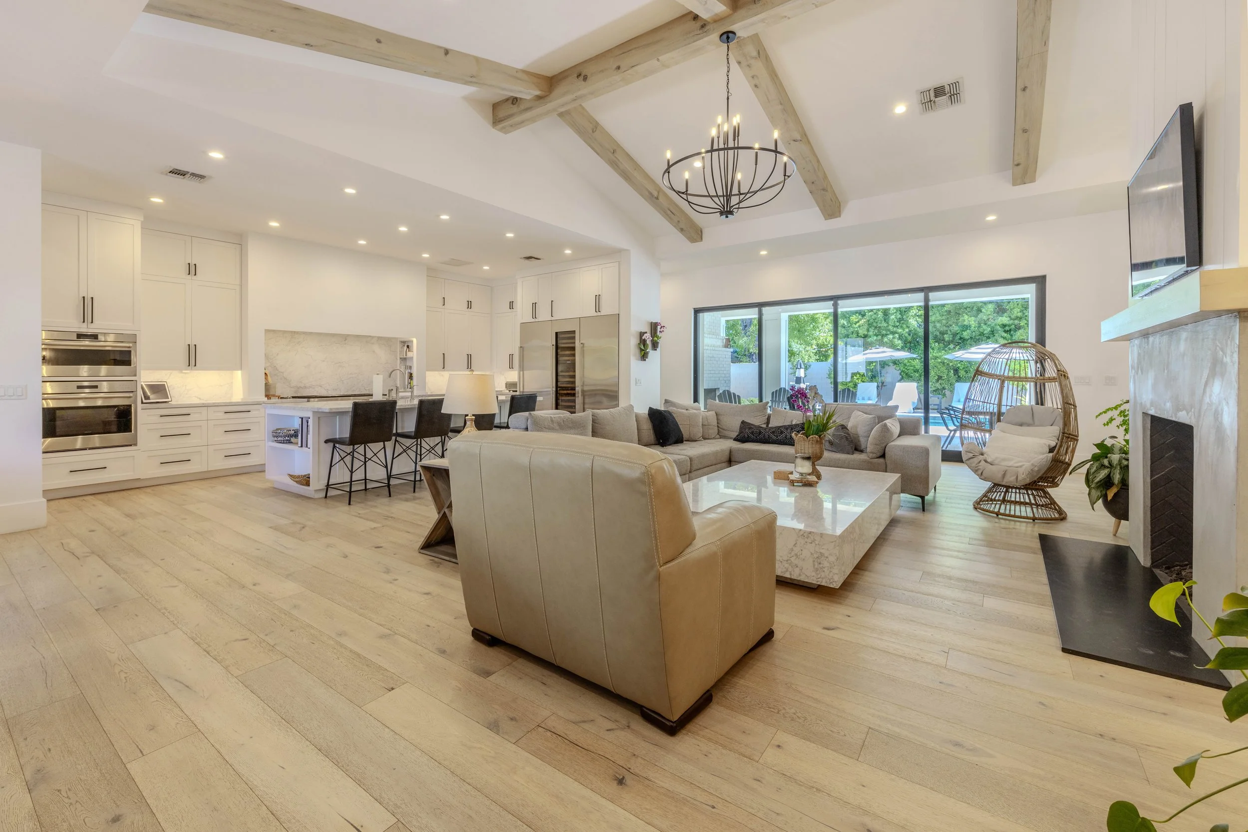 Modern open living room and kitchen with light wood floors, beige leather chair, large sectional couch, marble coffee table, and kitchen island with bar stools. Vaulted ceiling with exposed beams and chandelier. Sliding glass doors lead to outdoor pa
