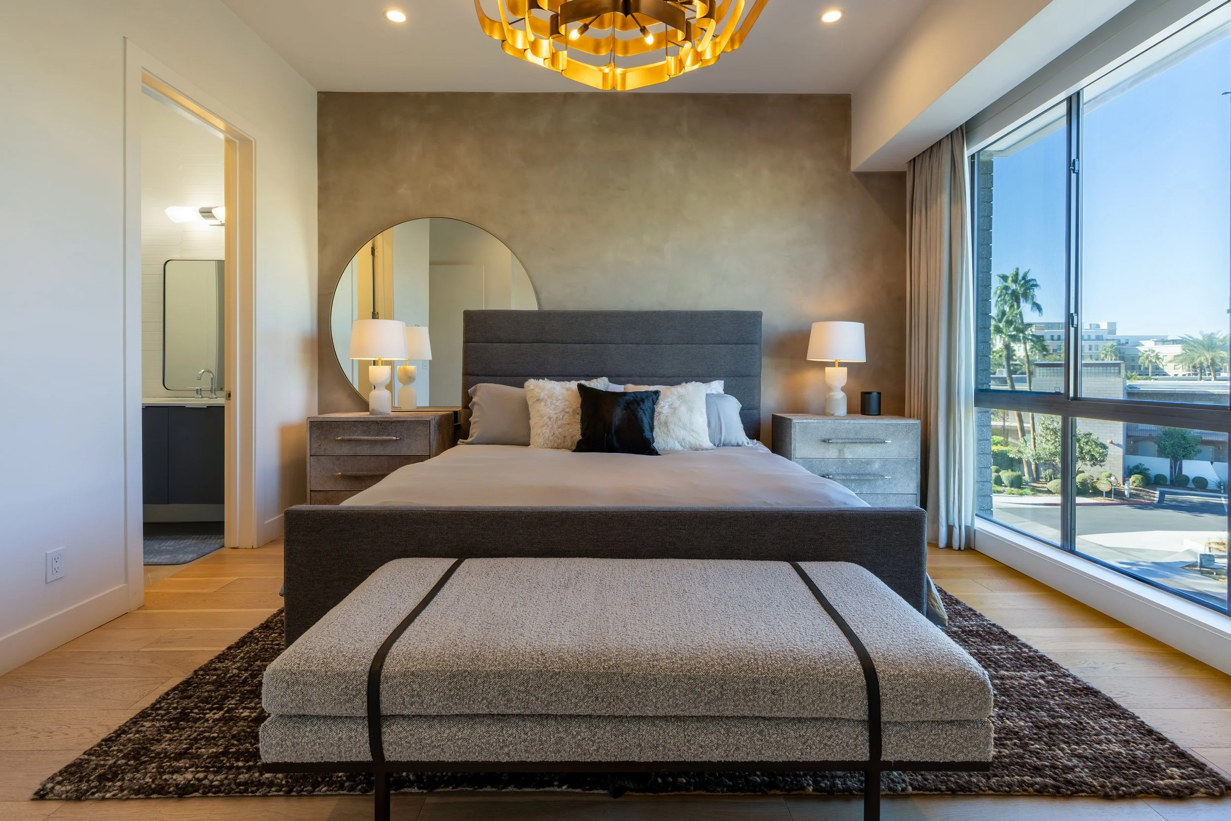 Modern bedroom with gray bed, side tables, lamps, large mirror, bench, and large window.