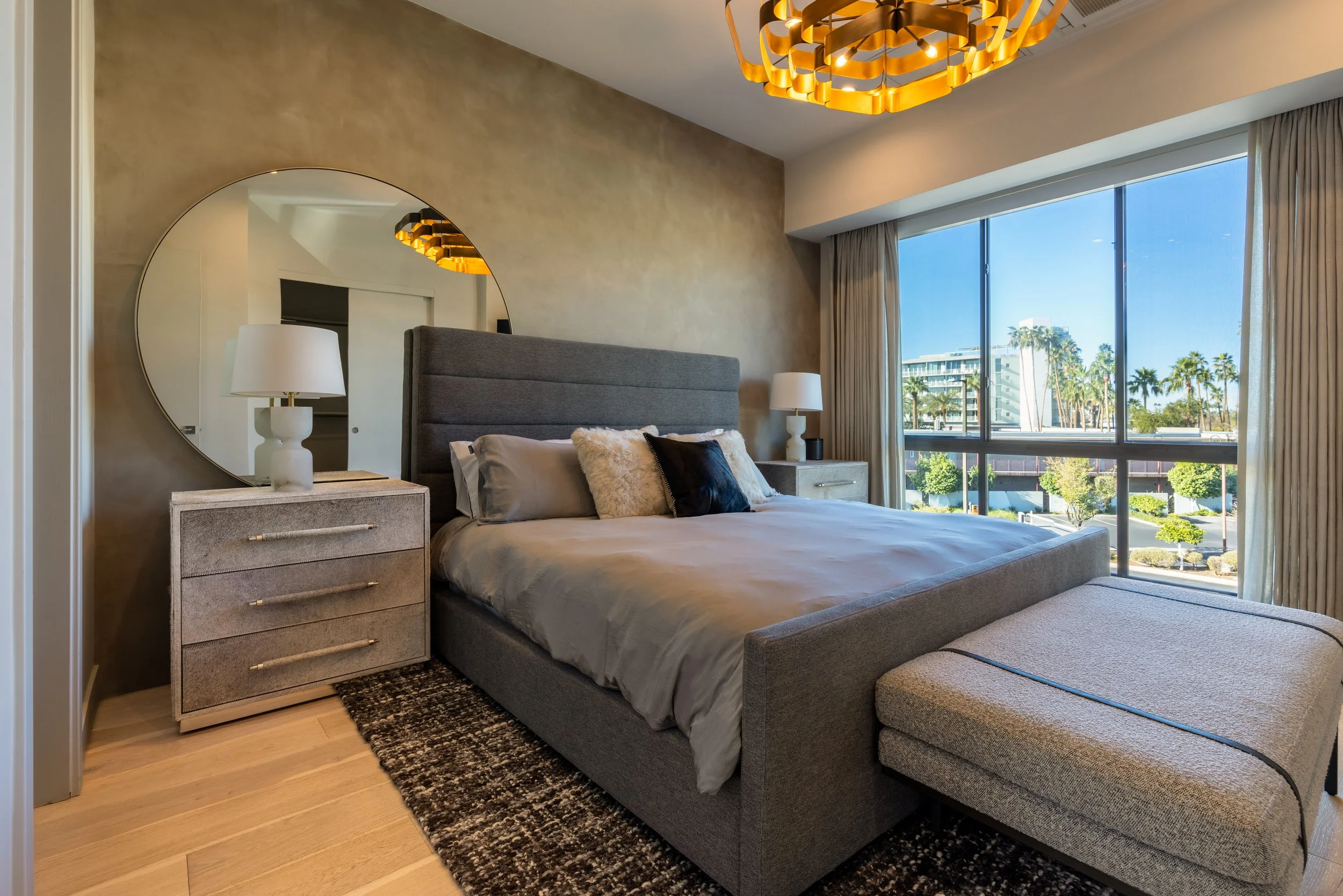 Modern bedroom with a large gray bed, round wall mirror, bedside tables with lamps, textured rug, and large window with city view.