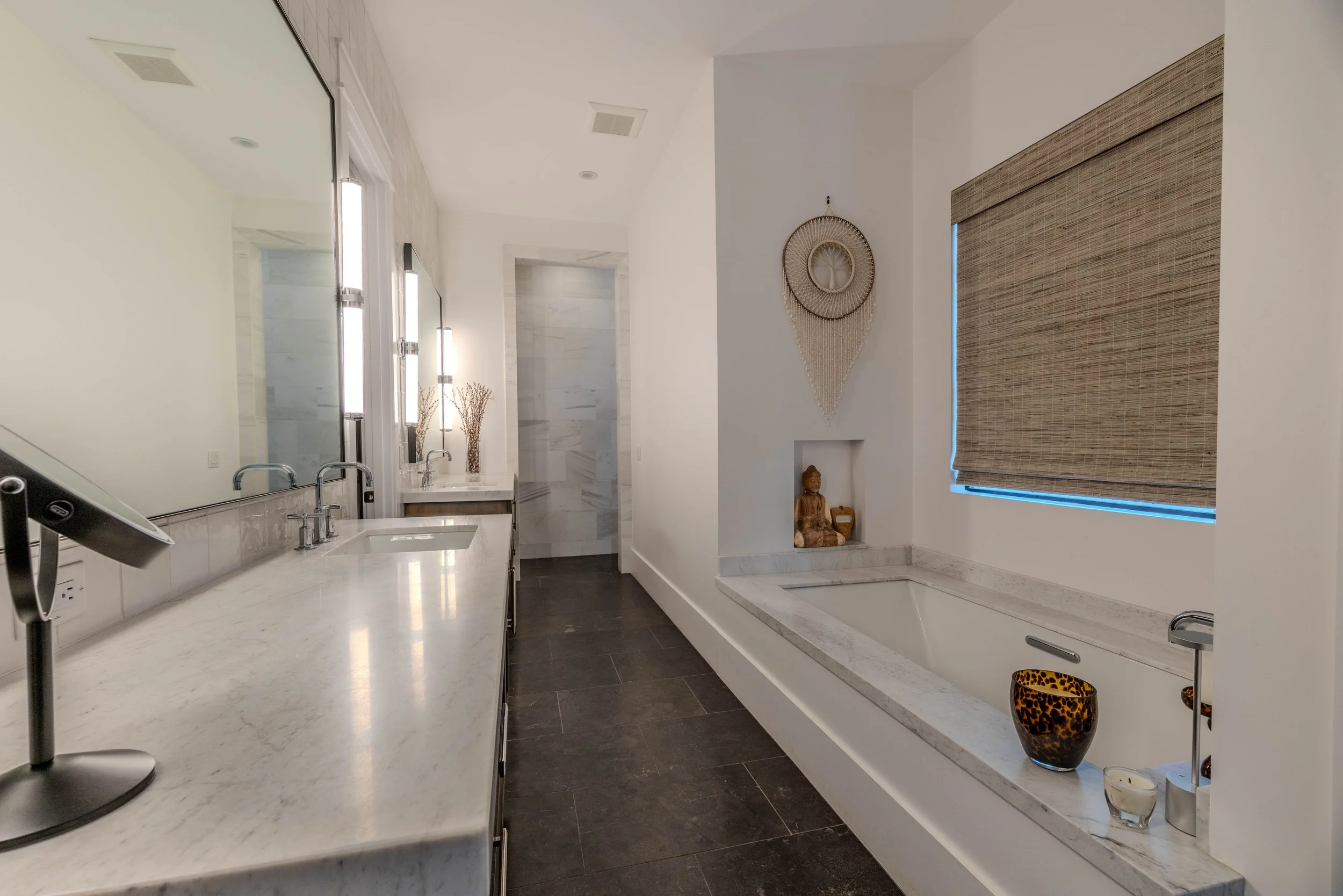 Modern bathroom with a marble countertop, large mirror, built-in bathtub, decorative items, and bamboo blinds.