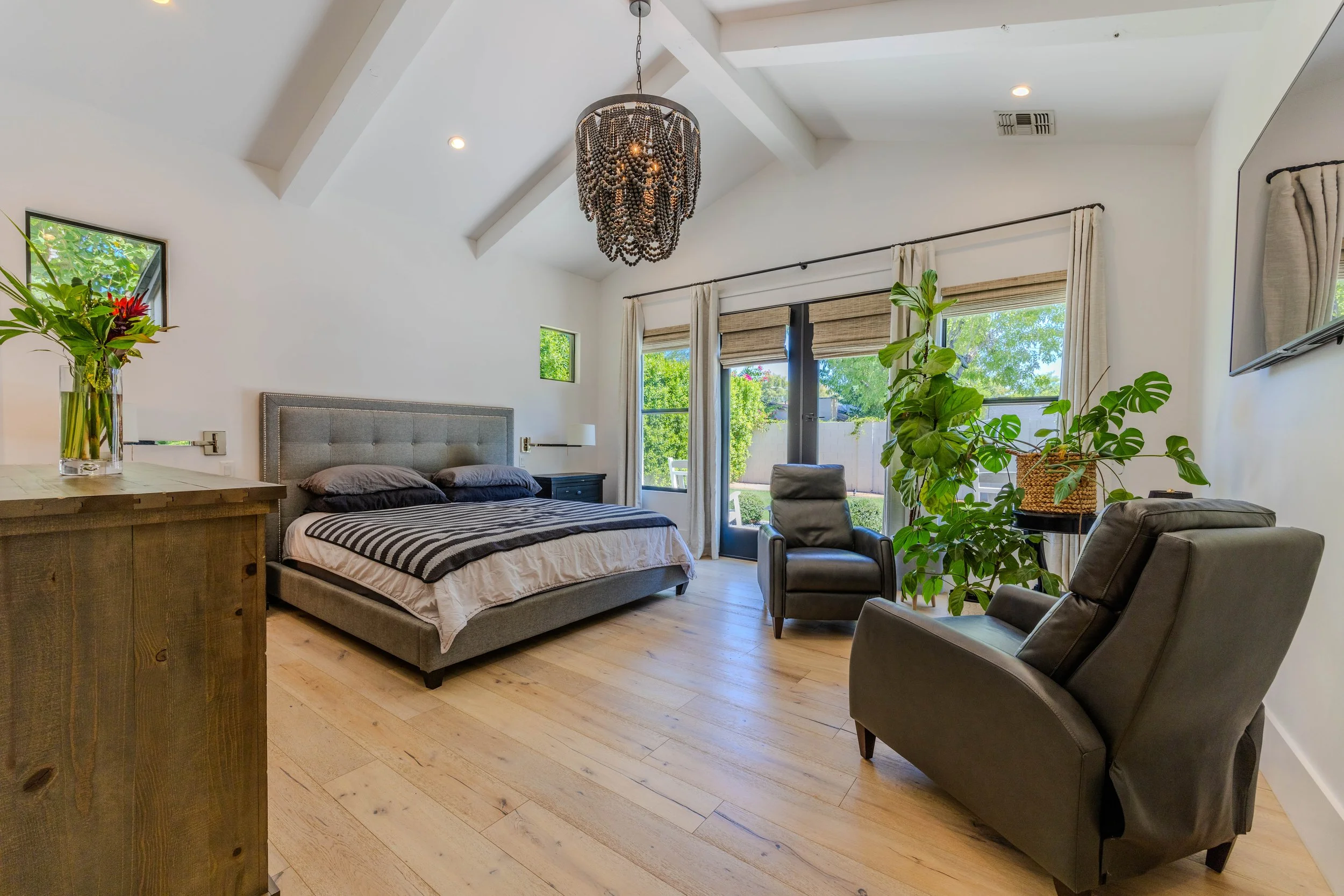 Modern bedroom with wooden flooring, featuring a gray upholstered bed, black and white bedding, armchairs, plants, a vase of flowers, and a large window with curtains.