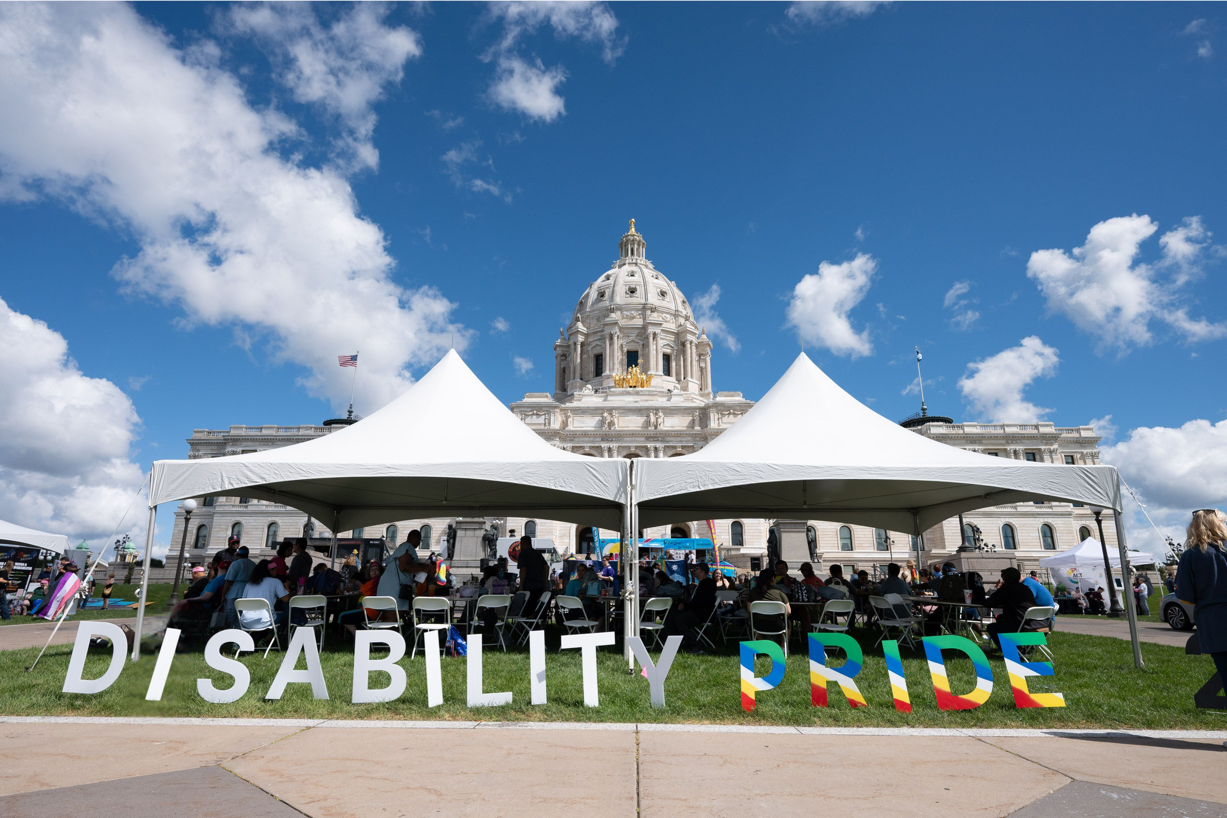 Minnesota State Capitol with blue sky. Tents are on the front lawn with the words 'Disability Pride' in front.
