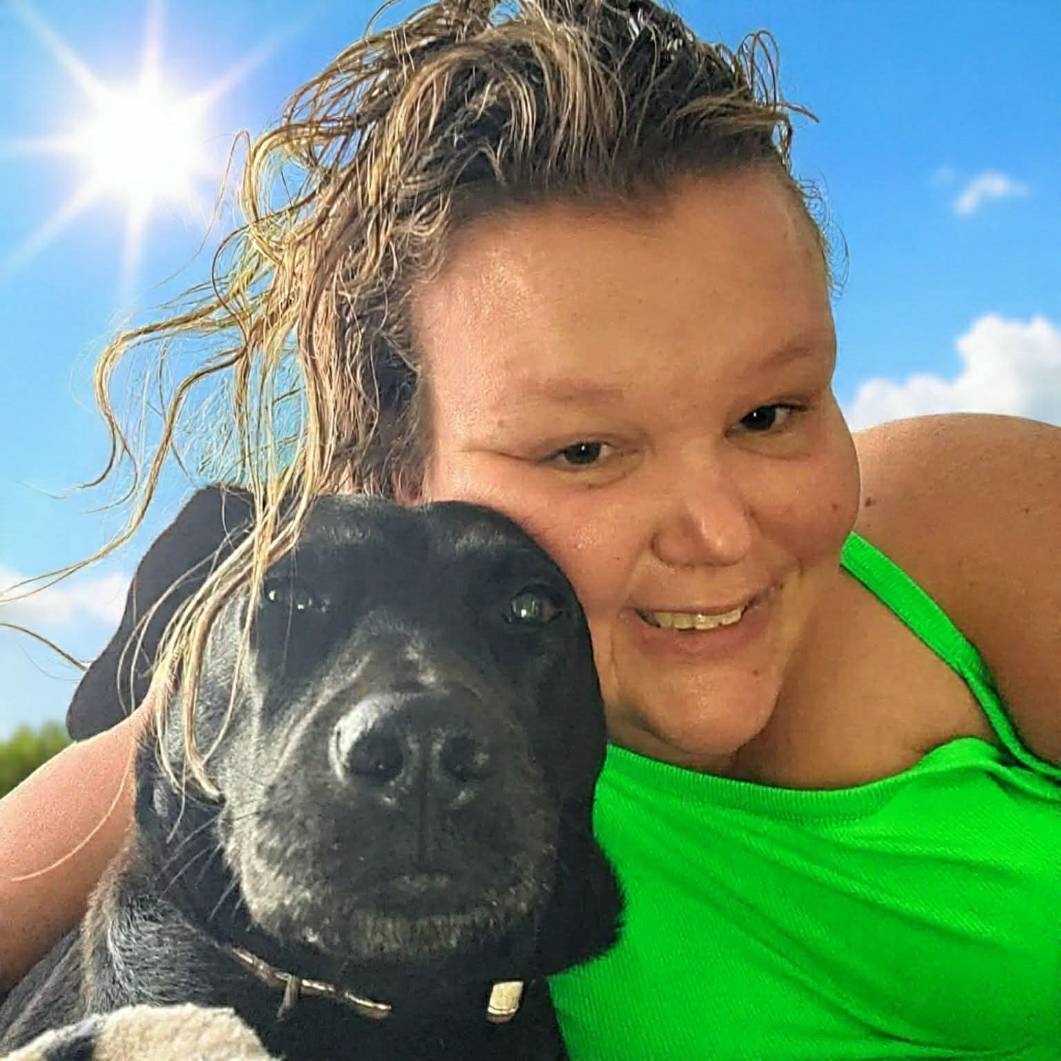 Carly smiling and wearing a green shirt in the sun. She is posing with a black dog.