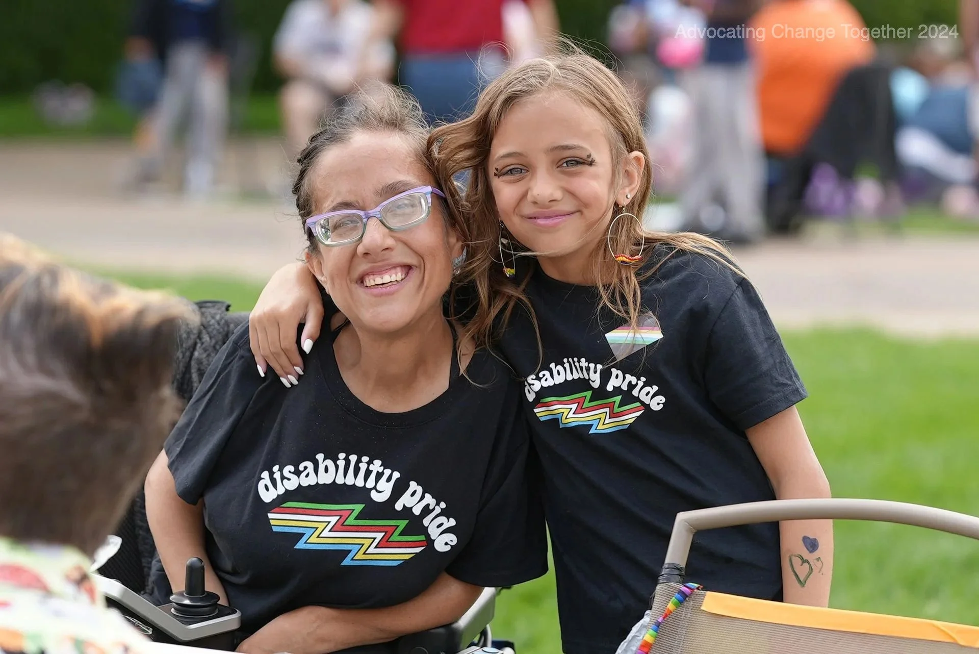 A woman and a younger girl wearing matching black disability pride t-shirts smile for a photo.