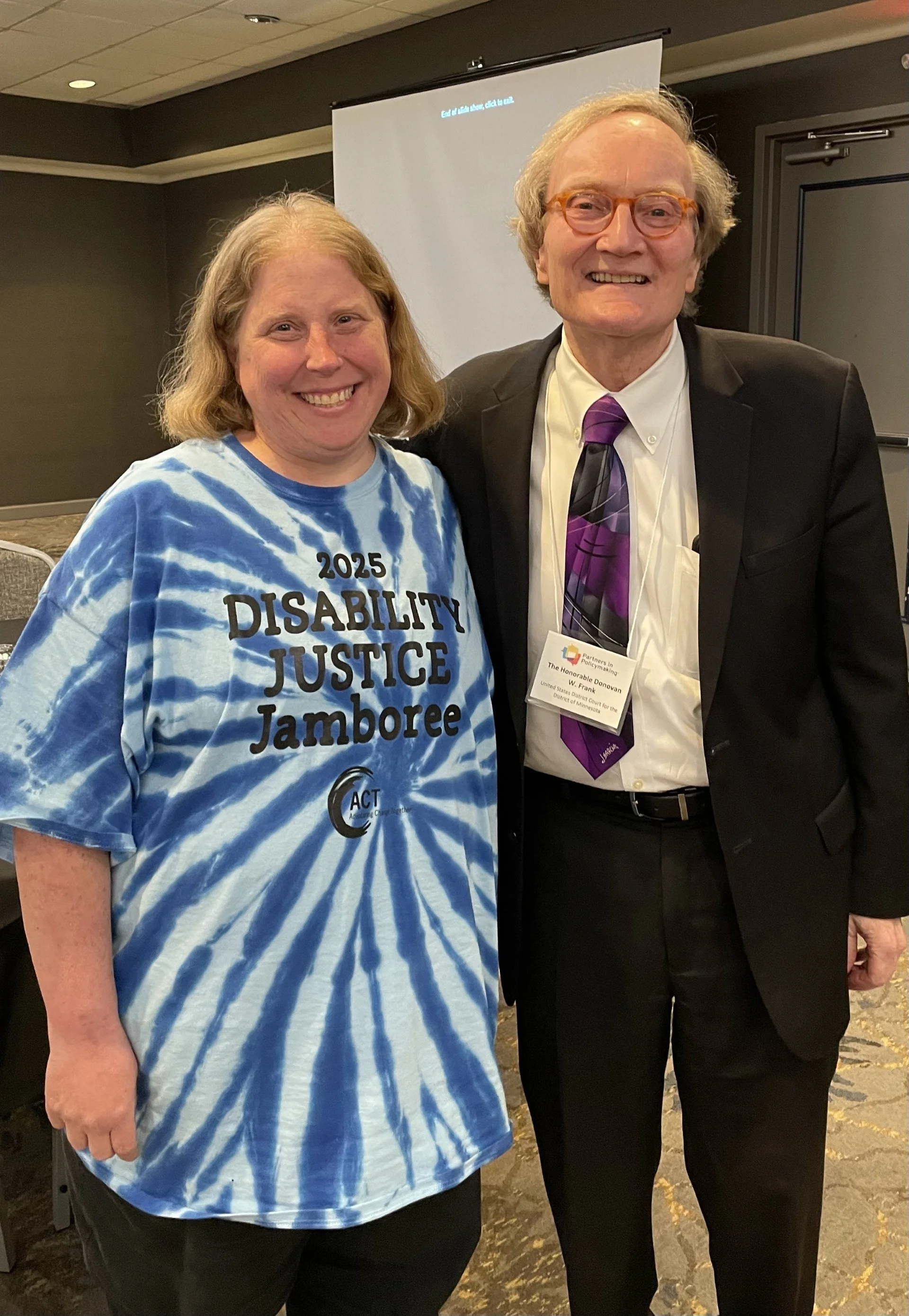 Jenny posing with Judge frank. Jenny is wearing a blue tie-dye shirt and Judge Frank is wearing  a suit and tie.