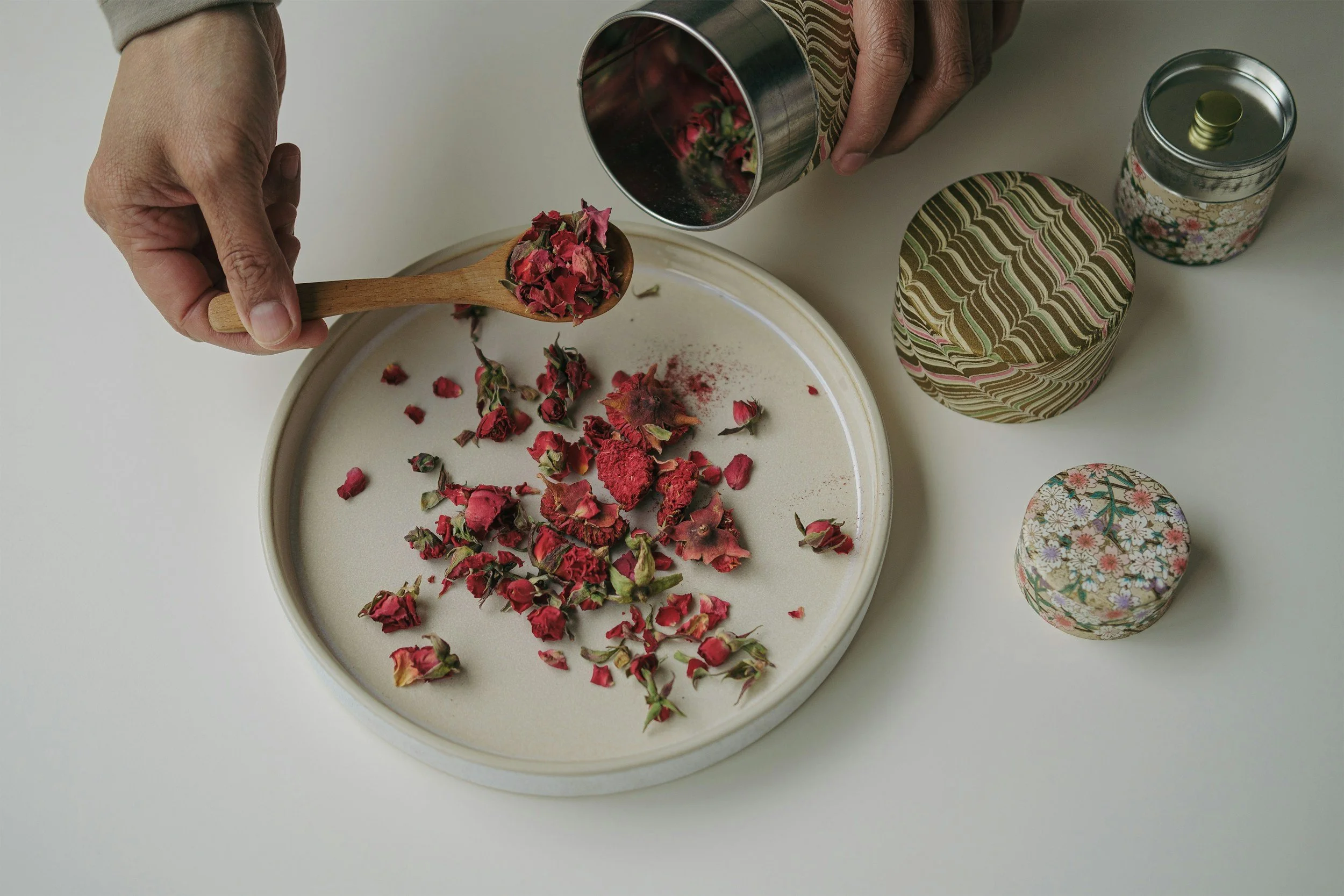 Person pouring dried rose petals onto a white tray, with decorated tins and jars nearby.