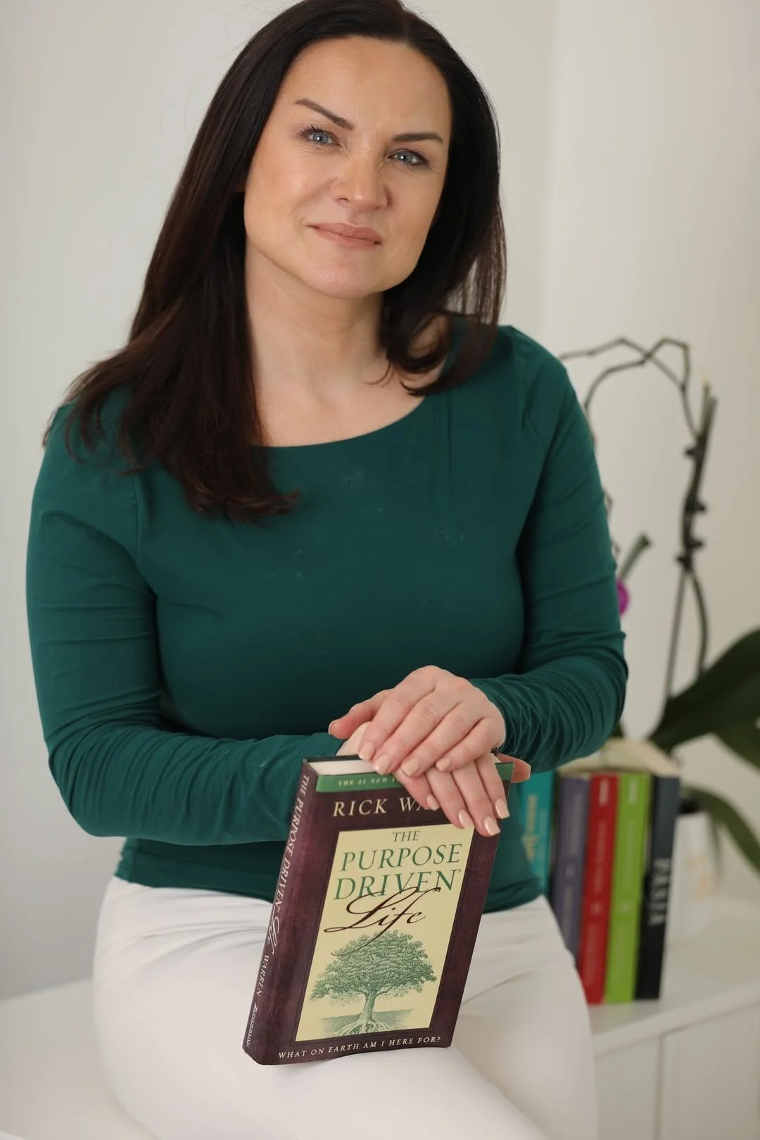 A woman in a green long-sleeve shirt holding a book titled 'The Purpose Driven Life.' She has dark brown hair and is sitting indoors with a small plant and a stack of books in the background.