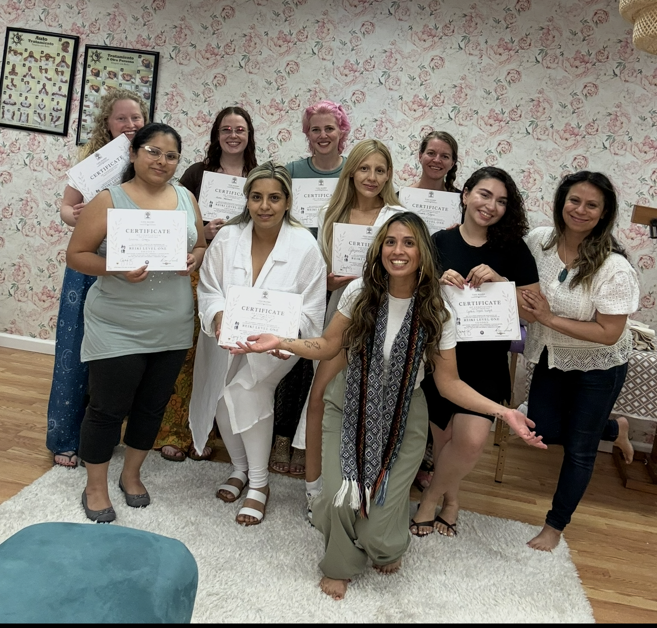 A group of ten women holding certificates and smiling, standing in a room with floral wallpaper, a wood floor, and a white rug. Some women are wearing casual clothing, and one has pink hair. A woman in the front is gesturing openly with her arms.