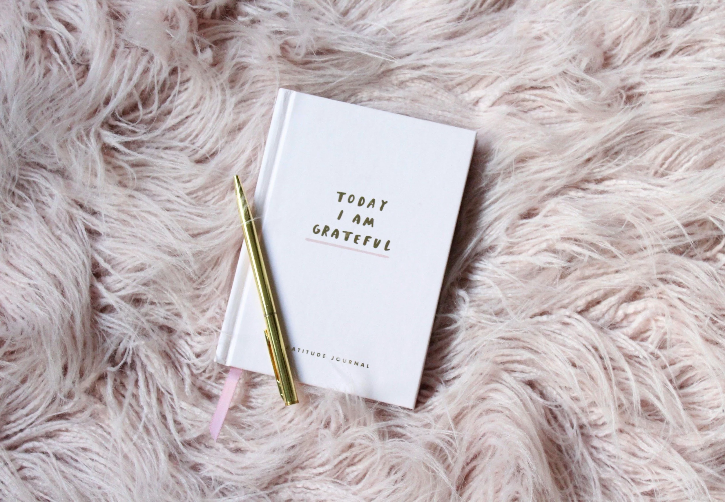 A white attitude journal with the words 'Today I am grateful' on the cover, a gold pen, and a pink ribbon bookmark, placed on a soft pink furry surface.