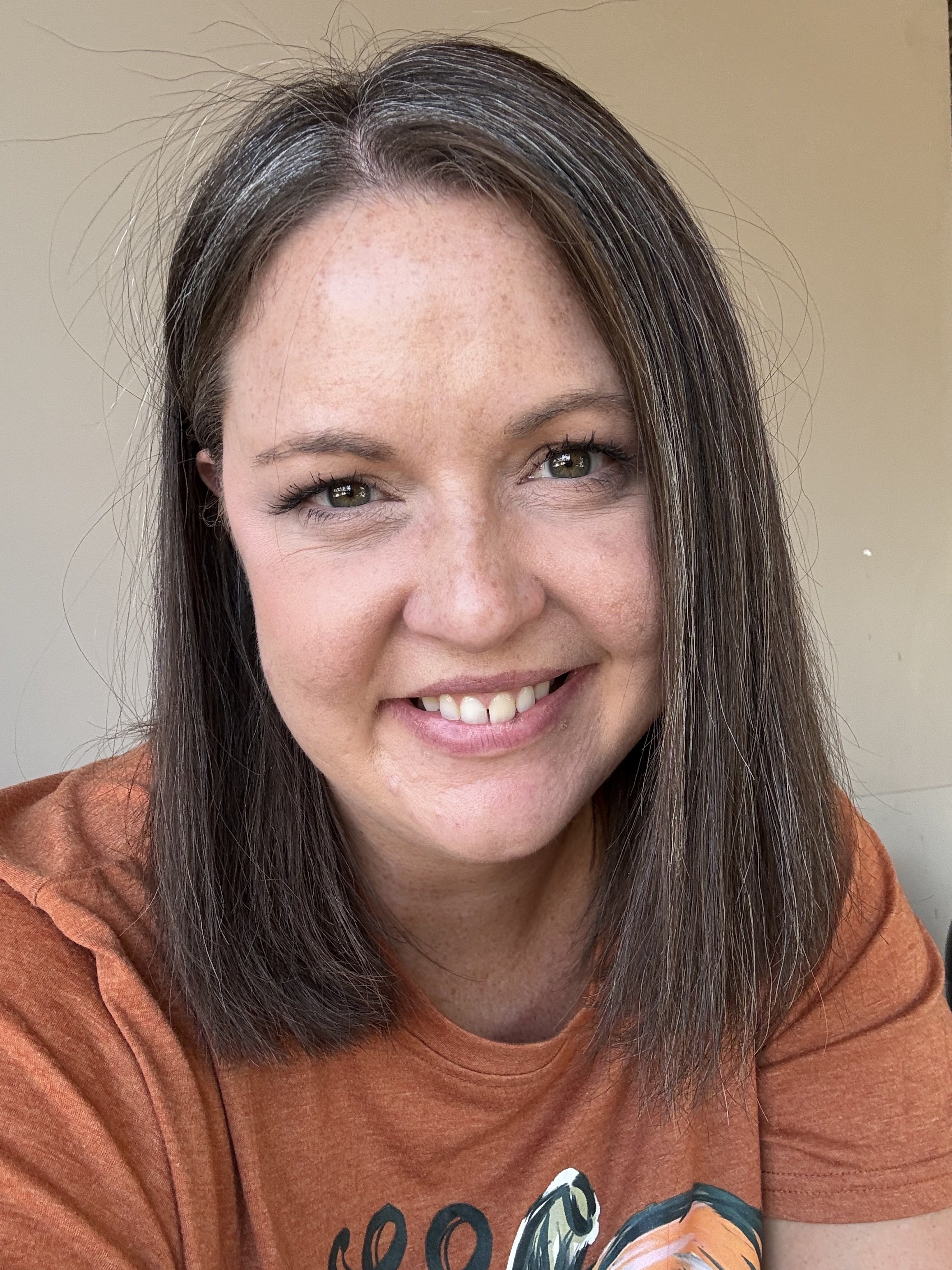 A close-up photo of a smiling woman with straight, shoulder-length brown hair and hazel eyes, wearing an orange shirt with a graphic design.
