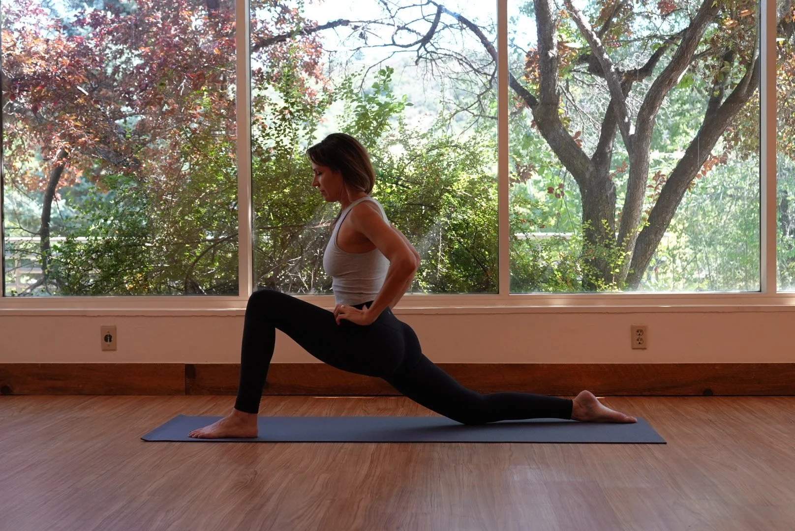 A woman practicing yoga indoors on a blue mat, doing a low lunge pose with a large window and green trees outside.