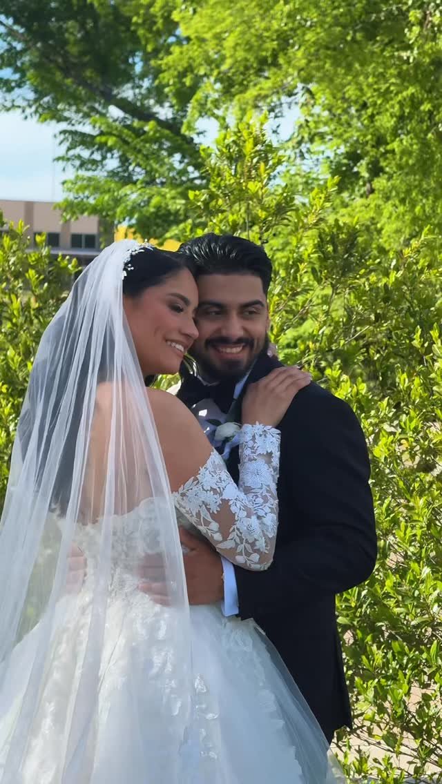 A bride and groom smiling together outdoors, with lush green trees and bushes in the background.