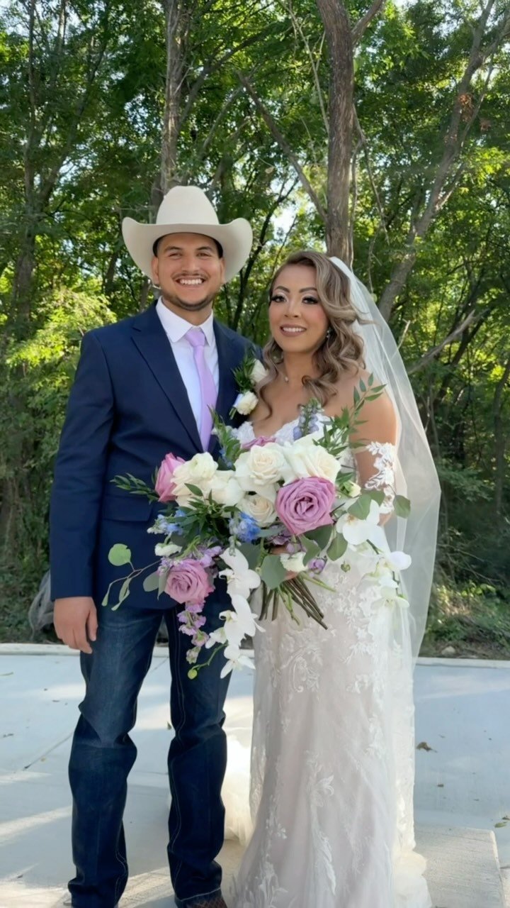 A bride and groom standing outdoors in a wooded area, smiling for a photo on their wedding day. The groom is wearing a dark blue suit with a white shirt, light pink tie, and a white cowboy hat. The bride is wearing a white lace wedding gown with a long veil, holding a large bouquet of white, pink, and purple flowers with greenery.