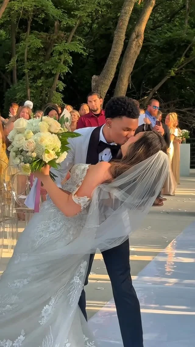 A newlywed couple sharing a kiss during their outdoor wedding ceremony, with guests in the background and trees in the setting.