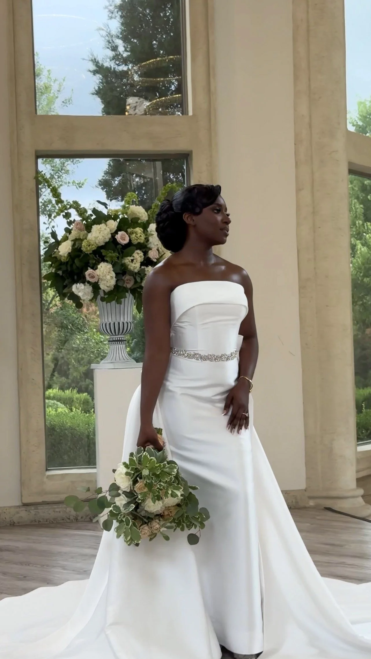 A woman in a strapless white wedding gown holding a bouquet of white and green flowers stands indoors near large windows with greenery outside.