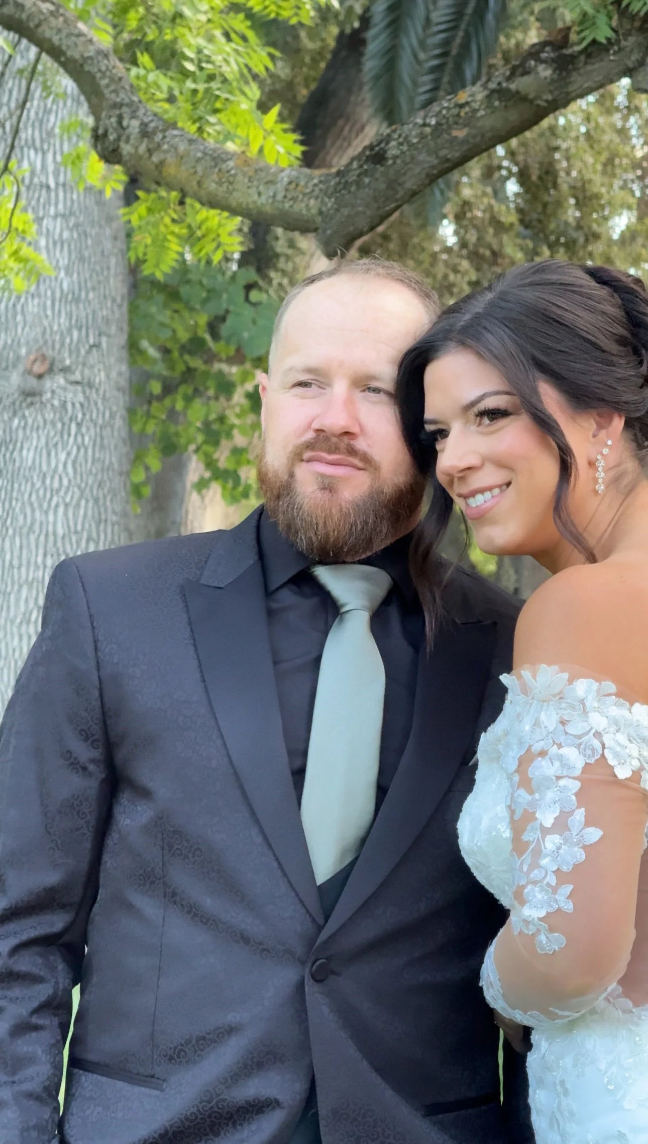 A newlywed couple taking a selfie outdoors. The groom is wearing a dark suit with a light tie, and the bride is in a white lace wedding dress with floral details and earrings. They are standing near a tree with green leaves, smiling and close to each other.