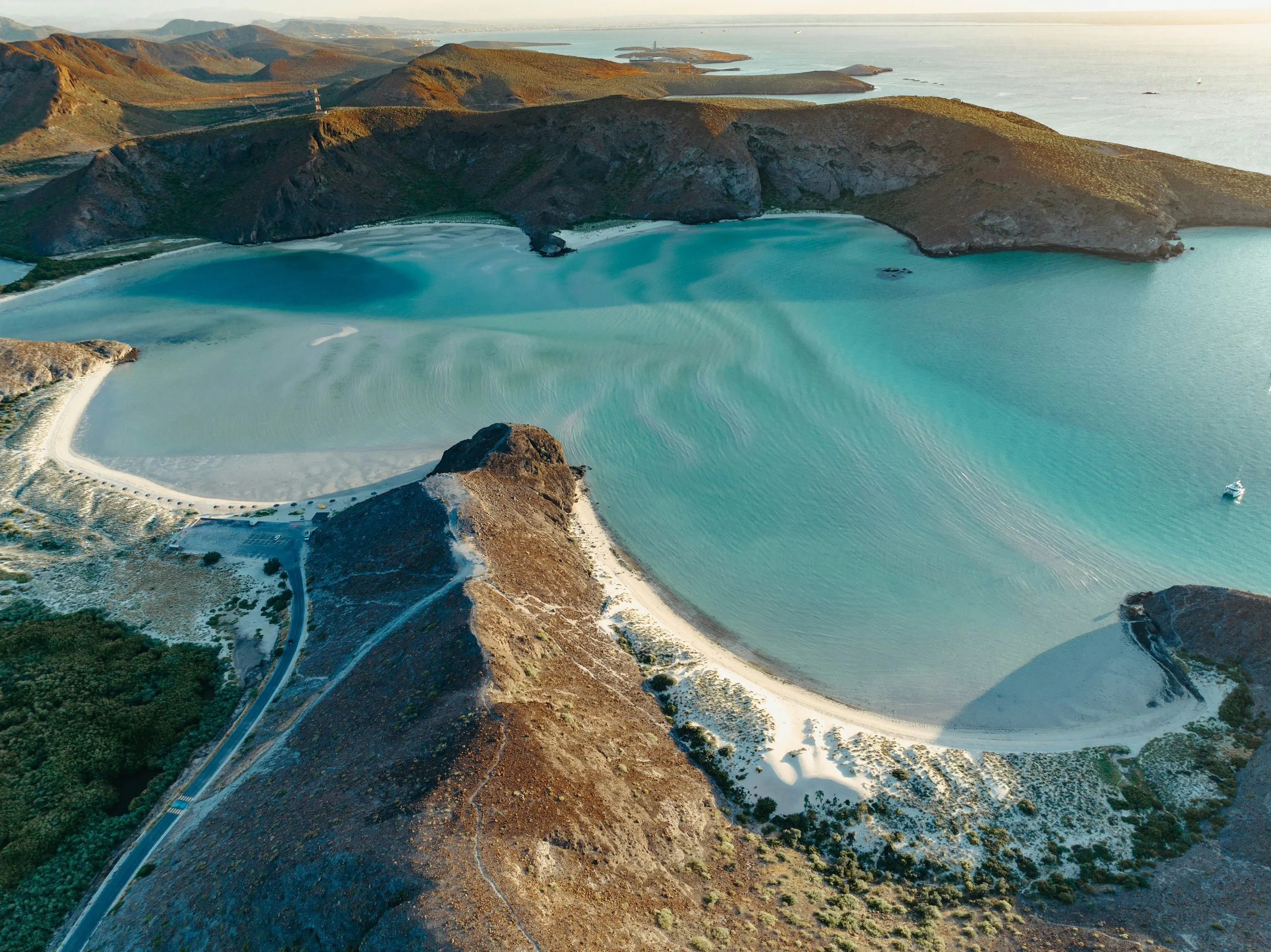 Vista aérea de una bahía con aguas turquesas, rodeada por colinas y montañas áridas, con una pequeña playa de arena blanca y un barco en el agua.